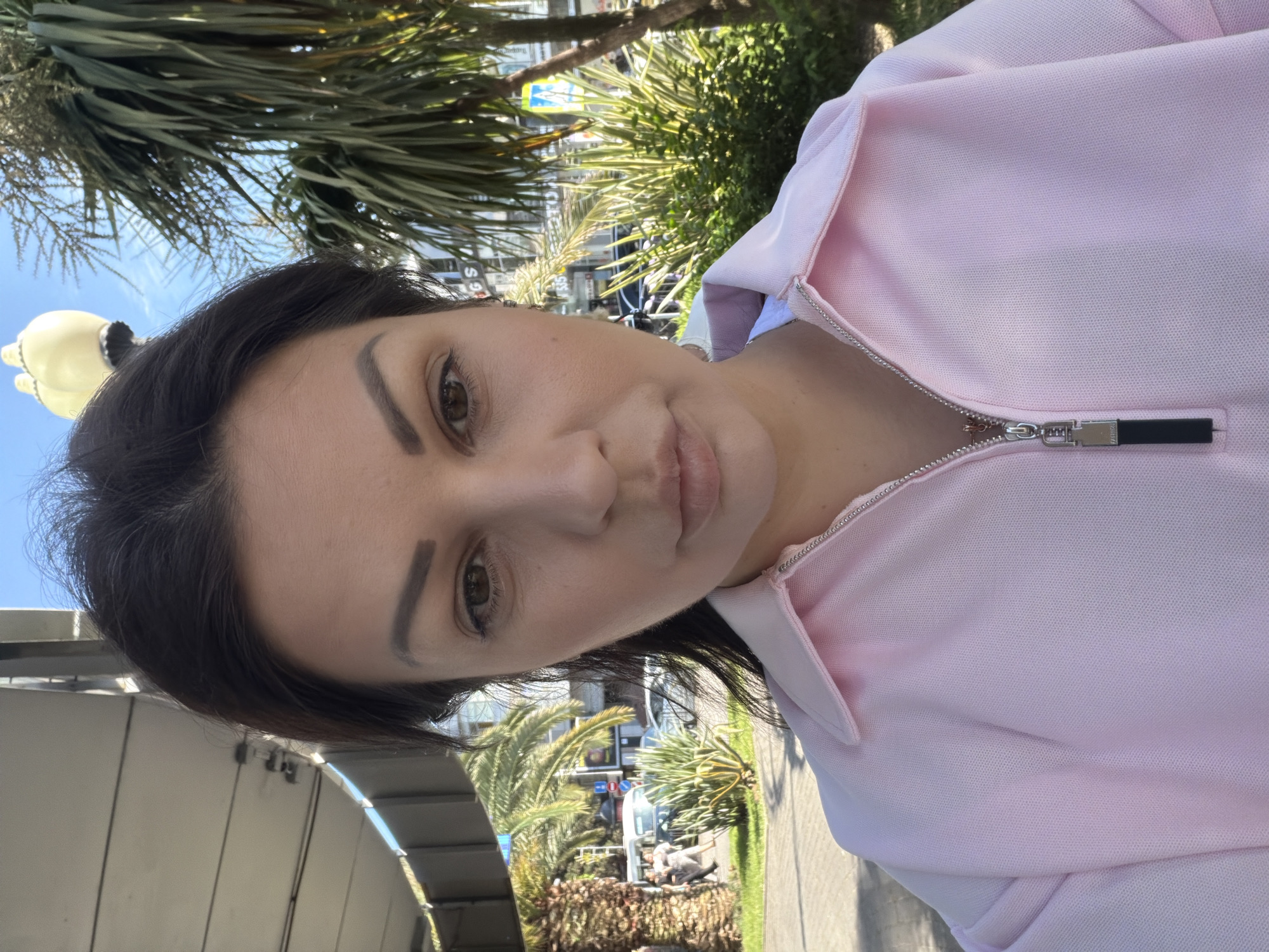 A close-up, over-the-shoulder selfie of a woman with dark hair and brown eyes, wearing a pink collared shirt with a zipper. She is outdoors in a sunny urban environment, with palm trees and buildings visible in the background. The location is the city of Sochi, Russia, possibly near a street or public walkway given the presence of a sidewalk, pedestrian crossing, and vehicles. The lighting suggests it is daytime.