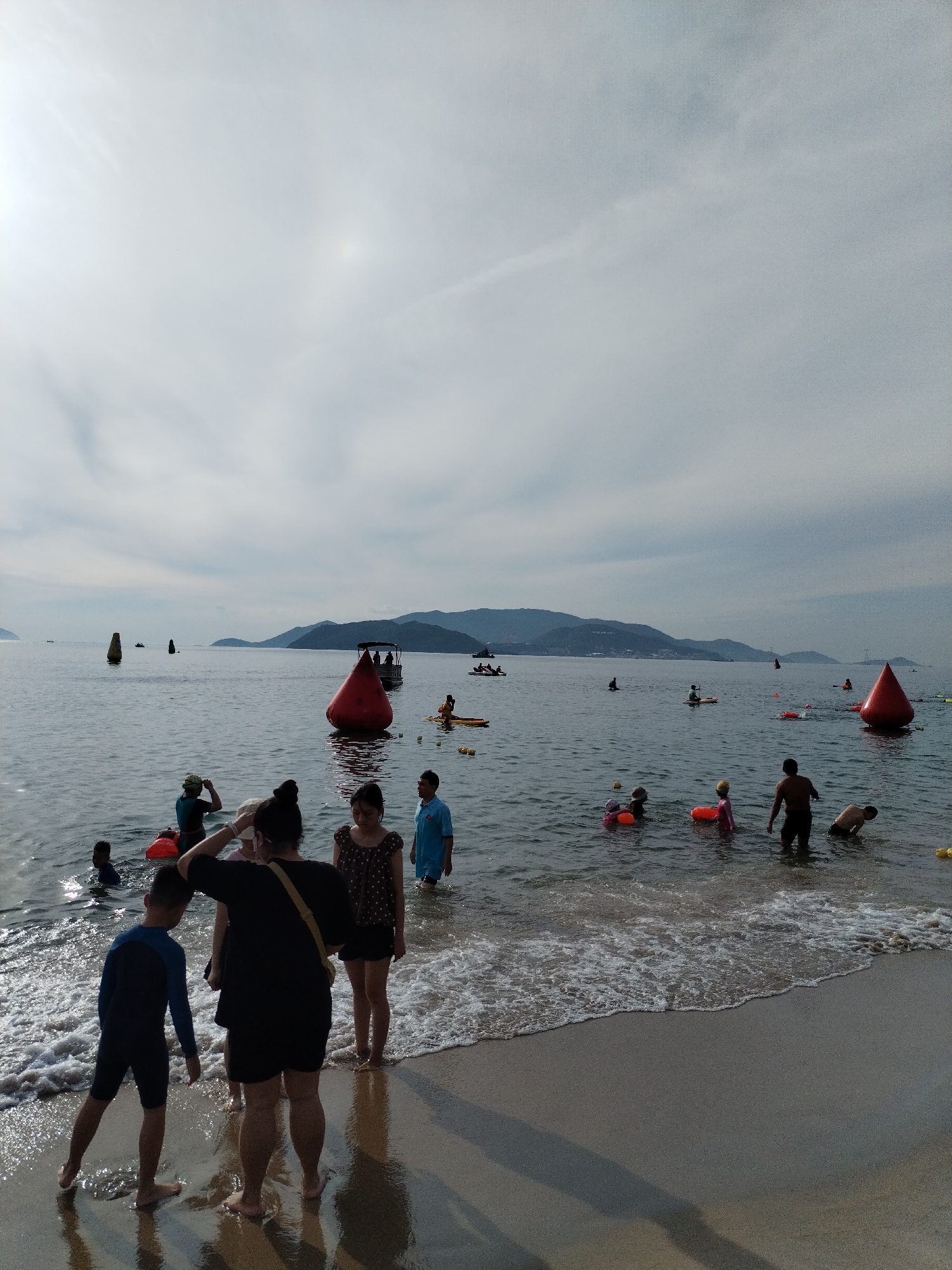 The image shows a beach scene in Nha Trang, Vietnam. The sky is overcast with the sun peeking through, creating a bright area in the upper left and a faint halo effect. The sea is calm with gentle waves lapping the shore. Several people are in the water, including children and adults. Some are standing, while others are swimming or playing.
In the foreground, a young boy in a blue and black rash guard stands on the wet sand, facing away from the camera. To his right, a woman in a black top and shorts stands with her back to the camera, her hand touching her head. Beside her, another woman in a polka dot top and shorts stands, looking to the right. To the far right, a man in a blue shirt and shorts stands in the shallow water.
In the middle ground, several red buoys are visible in the sea, marking a course or area. A boat with people on it is positioned behind a large red buoy. Other people are seen further out in the water, some on paddleboards. In the background, a range of hills or mountains forms the horizon.
The beach itself is sandy and wet from the incoming tide. The water appears clear with some foam from the waves. The overall impression is of a leisurely day at the beach with activities in the water.