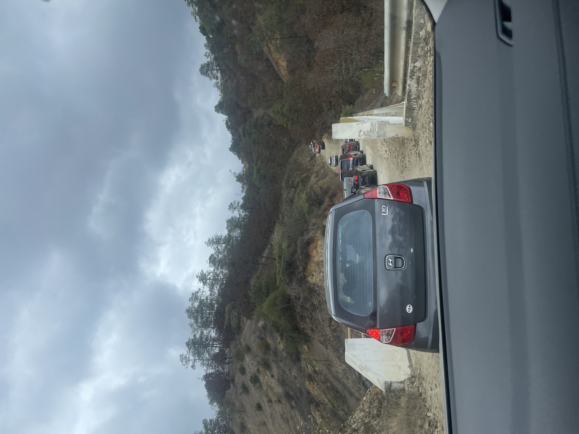The image shows a traffic jam on a narrow, dirt road in a mountainous area. The road is bordered by a steep hillside covered in trees on one side and a low concrete barrier on the other. Several cars are visible in the queue ahead, and the rear of a gray Hyundai i10 is prominently displayed in the foreground. The sky is overcast, suggesting a cloudy day. The road appears to be in La Cieneguilla, Mexico, based on the environment and type of landscape.