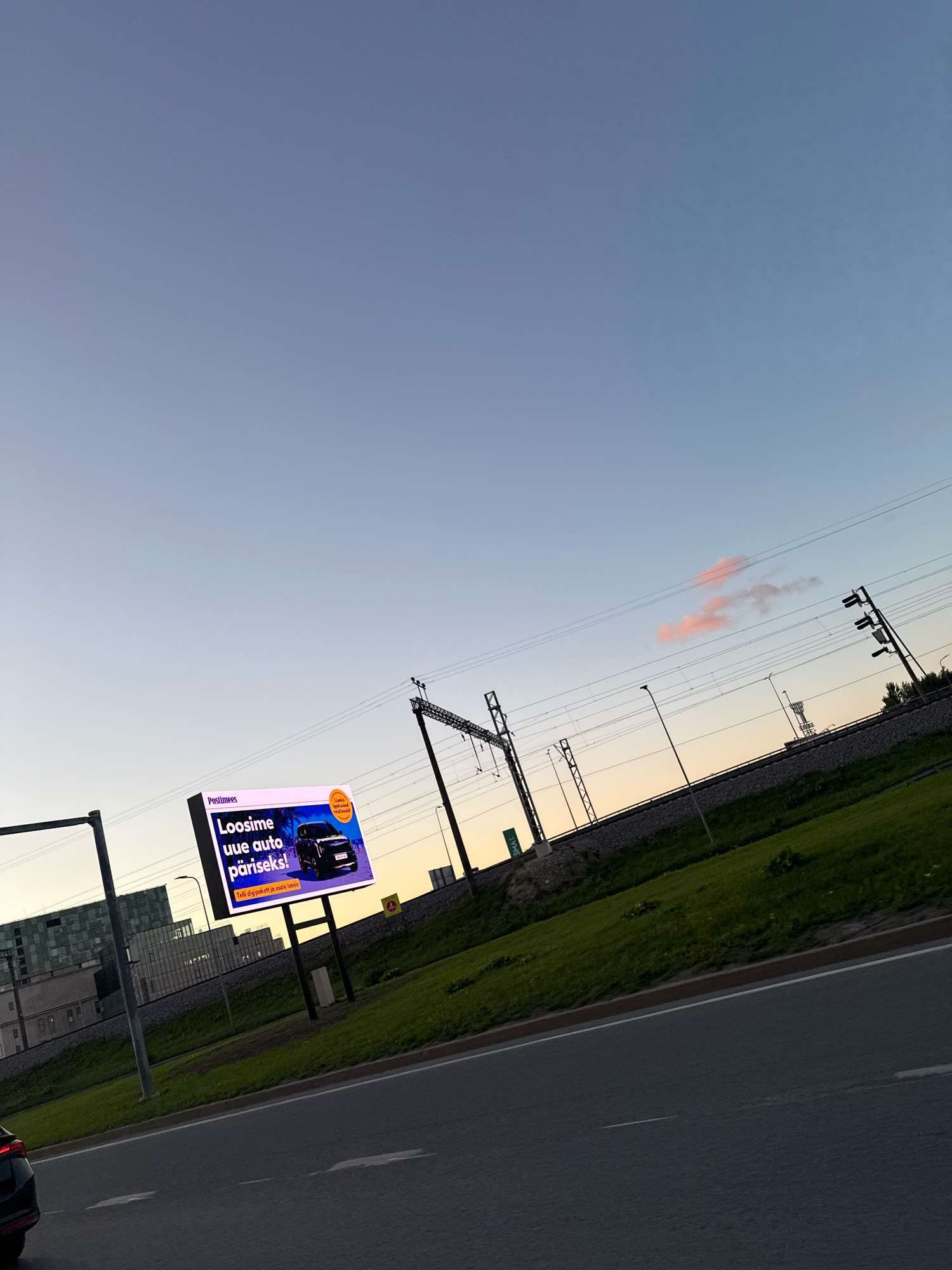 The image shows a digital billboard advertising a car giveaway. The billboard, which is white with purple and orange accents, displays the text "Loosime uue auto päris!". There's an image of a black SUV on the right side of the billboard. The billboard is located in Tallinn, Estonia, near a set of train tracks and a highway. The sky is a pale blue with some faint pink clouds. There are power lines and streetlights visible. The time appears to be either sunrise or sunset.