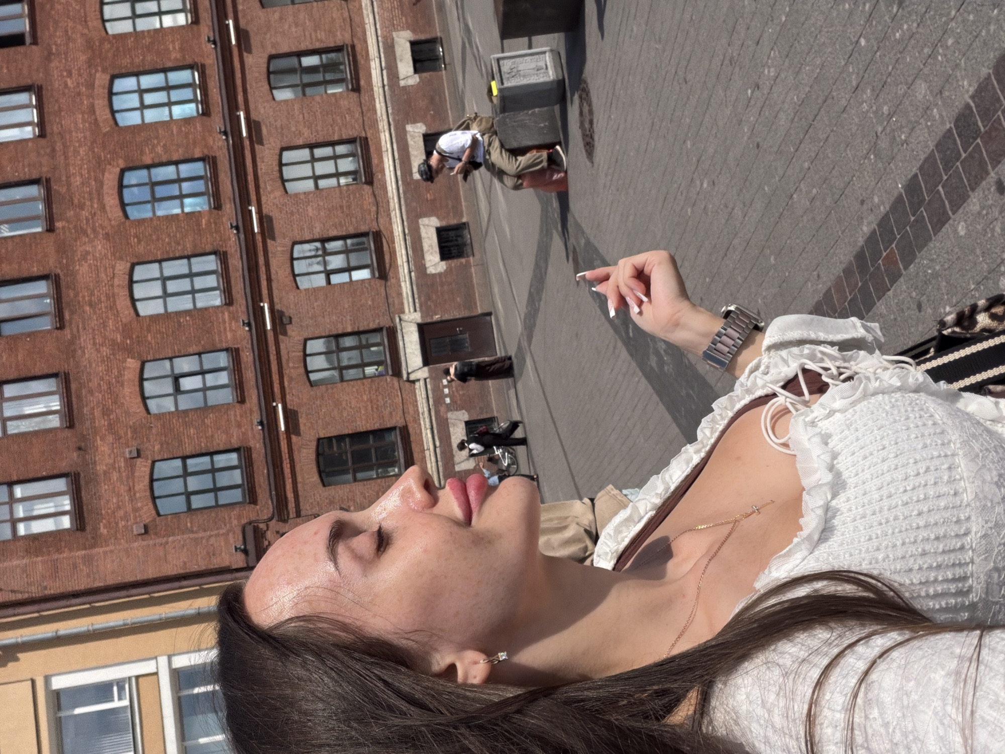 The image captures a close-up of a young woman's face and upper body, shot from a low angle. She is outdoors, with the sun casting strong shadows, suggesting it is a bright, sunny day. Her head is tilted back, eyes closed, and lips slightly parted, conveying a sense of relaxation or enjoyment. She wears a white, ruffled top and a delicate gold necklace with a small cross pendant. A silver watch is visible on her wrist.
In the background, a multi-story brick building with numerous windows dominates the left side of the frame. To the right, a cobblestone street is visible, with a man in a white shirt and tan pants sitting on a large dark object. Further in the distance, other people and a bicycle can be seen. The overall setting appears to be an urban environment, possibly a public square or street in Saint Petersburg, Russia, given the architectural style of the building and the cobblestone pavement. The time of day is clearly daytime due to the strong sunlight and shadows.