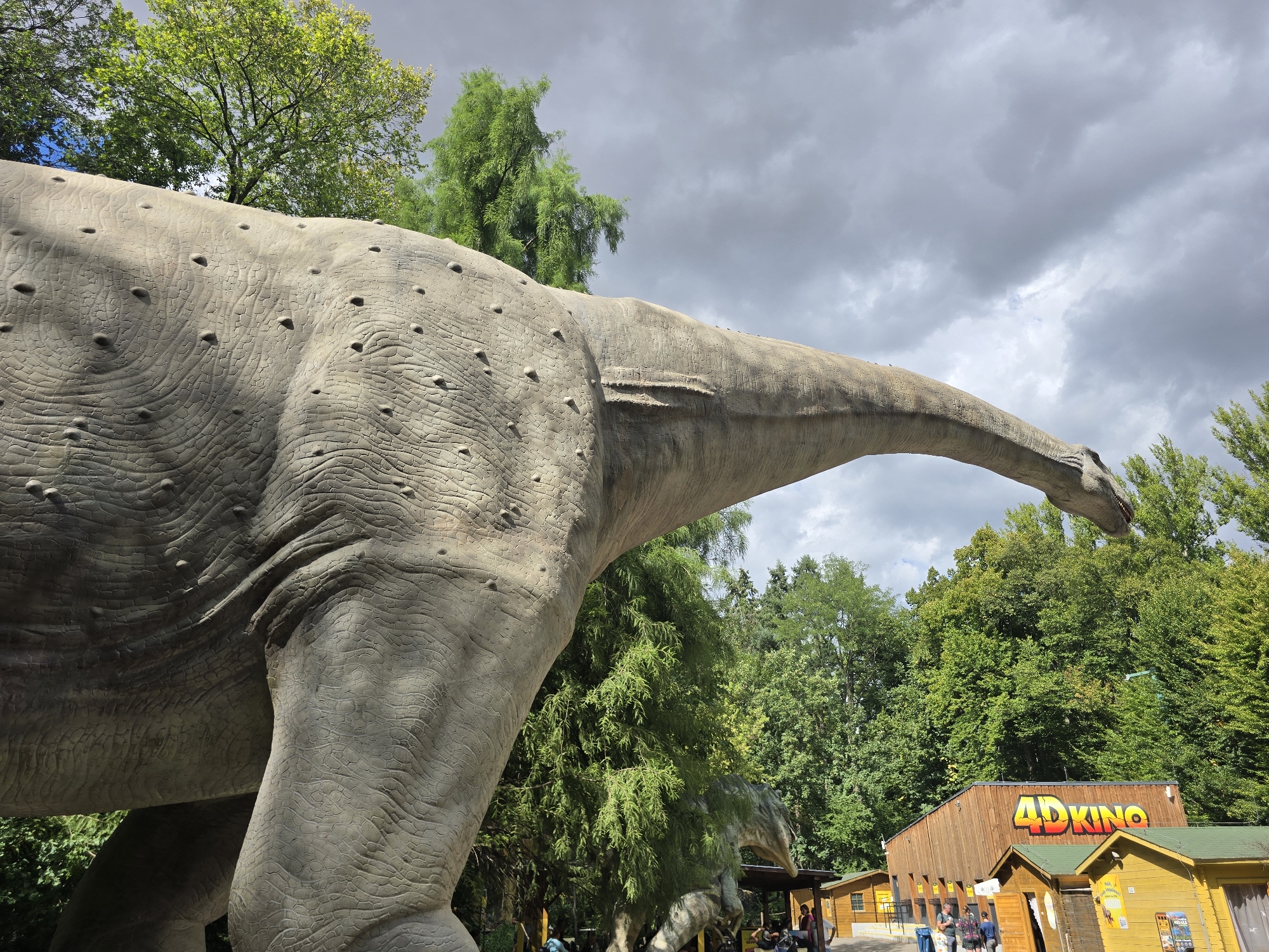 A life-sized replica of a dinosaur, likely a sauropod due to its long neck and tail, stands prominently in the foreground. The dinosaur model is textured to resemble rough, scaly skin with numerous small bumps. Its head and neck extend to the right, reaching towards a cloudy sky. Behind the dinosaur, a wooden building with "4DKINO" signage is visible, suggesting it's part of an attraction or amusement park. Several other smaller wooden structures are also present. In the background, lush green trees form a dense forest canopy, with some foliage appearing lighter green and yellow, possibly indicating sunlight filtering through or a mix of tree species. A few people are scattered around the area, near the buildings and pathways. The location is identified as Vyškov, Czechia. The overall scene depicts an outdoor attraction with a focus on dinosaur models.