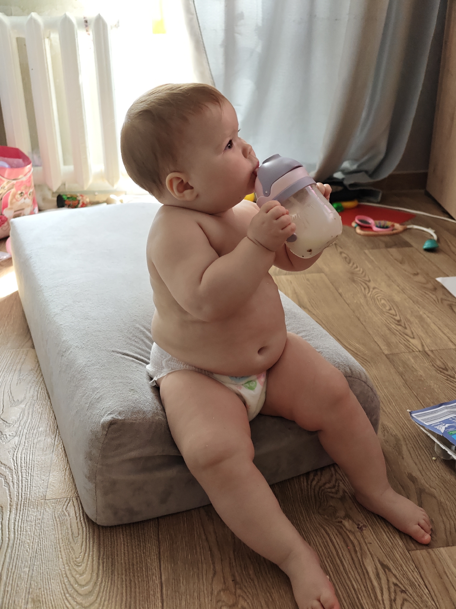 A baby is sitting on a grey cushion, drinking from a bottle. The baby is wearing a diaper. The location is Chaikovsky, Russia. The background includes a white radiator, a window with a curtain, and a wooden floor with various toys. There is a pink container in the background.