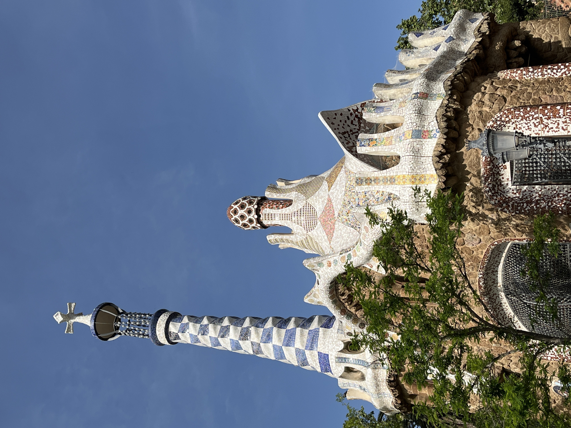 The image captures a distinctive architectural marvel against a clear blue sky, viewed from a low angle looking upwards. The primary subject is a building or structure characterized by its elaborate, mosaic-covered surfaces and organic, undulating forms, strongly indicative of Antoni Gaudí's work in Barcelona, specifically Park Güell.
The structure features an intricate roofline with wave-like contours, entirely adorned with colorful `trencadís` (broken tile mosaics). These mosaics showcase a vibrant array of patterns and hues, including white, blue, yellow, red, and brown, arranged in geometric and abstract designs.
A prominent, slender tower-like element rises towards the left, topped with a simple white cross. This tower is covered in a striking blue and white checkerboard mosaic pattern. Further to the right, another dome-shaped or bulbous architectural feature, also covered in mosaics (reddish-brown and white), adds to the whimsical character of the building.
Parts of the building appear to be constructed or integrated with rough-hewn stone or rock, creating a textural contrast with the smooth, detailed mosaics. Lush green foliage from trees partially frames the structure on the right side and lower parts, suggesting a garden or park setting.
The scene is set outdoors under a bright, cloudless blue sky, indicating clear, sunny weather during the daytime, likely morning or early afternoon due to the soft yet vibrant illumination. There are no people visible, and no discernible text appears on the structure. The overall impression is one of intricate artistry and unique design within a natural landscape.