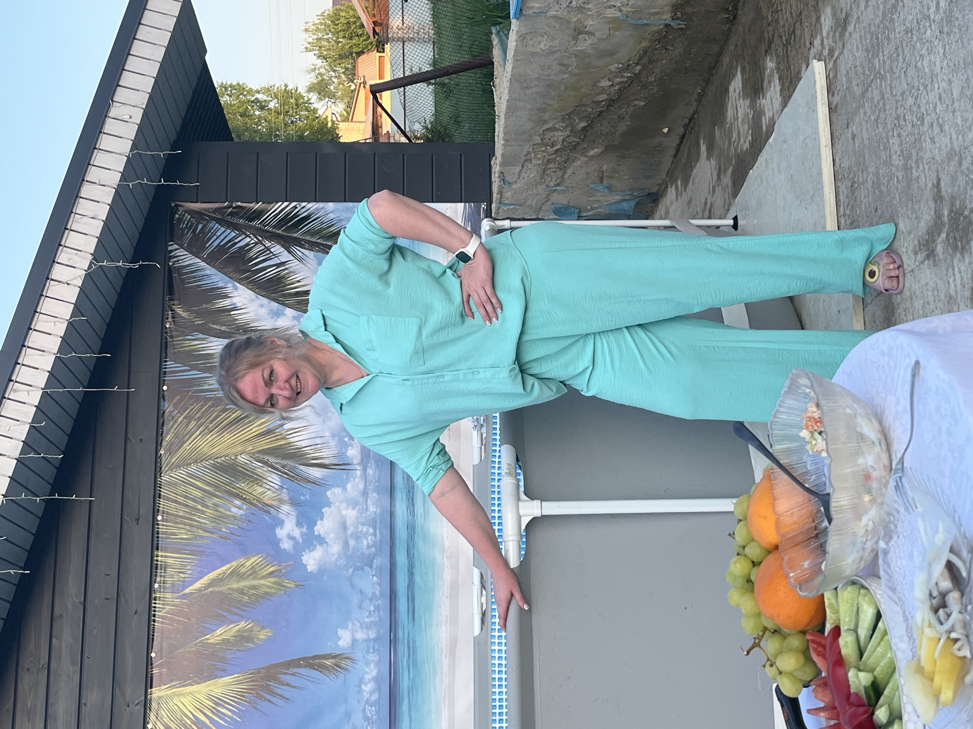 The image depicts a woman standing near what appears to be an above-ground pool, likely in a backyard setting. The location is identified as Новая Чернушка, Russia. The woman is dressed in a light blue pantsuit. Behind her is a backdrop featuring a beach scene with palm trees and a sky, attached to what looks like a shed or building with dark siding. A table with food, including fruit and salad, is in the foreground. The weather appears to be clear and sunny, based on the lighting.