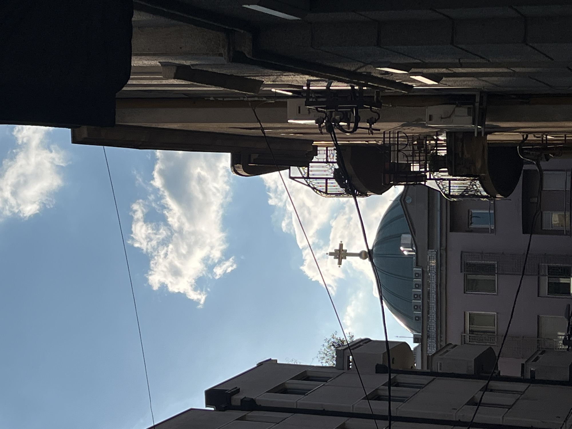 The image shows an upward-angled view in Belgrade, Serbia. A dome of an Orthodox Christian church with a cross is visible against a partly cloudy blue sky. Buildings are present in the foreground and background. Utility lines crisscross the scene. Balconies and air conditioning units are visible on one of the buildings. The perspective is from a narrow street or alley, with building overhangs visible at the top of the frame.
