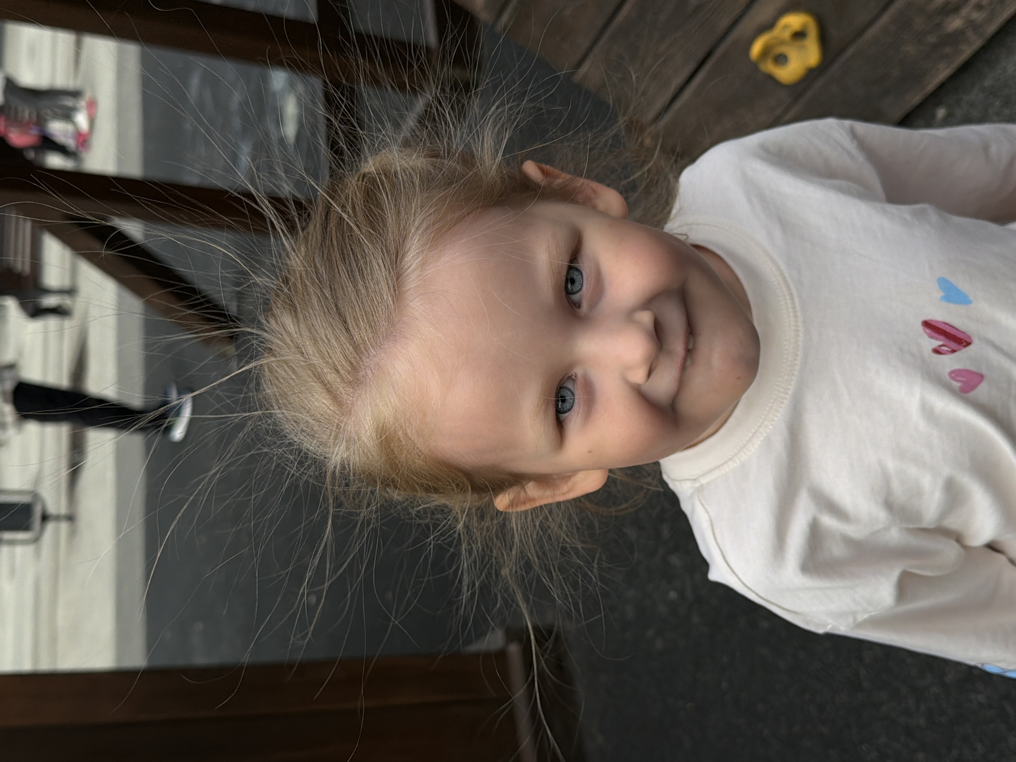 A young, blonde-haired child with blue eyes is angled in an outdoor setting. The child is wearing a white shirt with pink and blue heart designs. Her hair is displaying static electricity, standing out from her head. A wooden structure and a dark, possibly paved surface surround the child. A portion of what appears to be playground equipment, specifically a yellow climbing hold, is visible in the background. The child is looking directly at the camera. There are no identifiable features to place this in the city of Saratov, Russia.