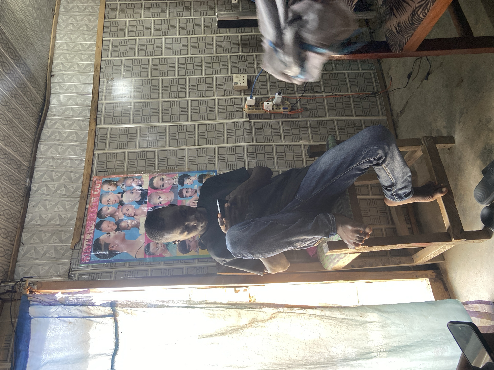 This image depicts a person sitting on a wooden structure, looking at their phone. The person is wearing blue jeans and a dark top. They are positioned in what appears to be a room with patterned wallpaper. On the wall, there is a poster for "SISTERS HAIR CUT" featuring various hairstyles. Electrical outlets and extension cords are also visible on the wall. The scene is illuminated by natural light coming from a window covered with translucent material, suggesting it is daytime. The overall impression is of a casual indoor setting, possibly a home or a small business, in Kuson, Nigeria. The person's focus on their phone suggests engagement with digital content or communication.