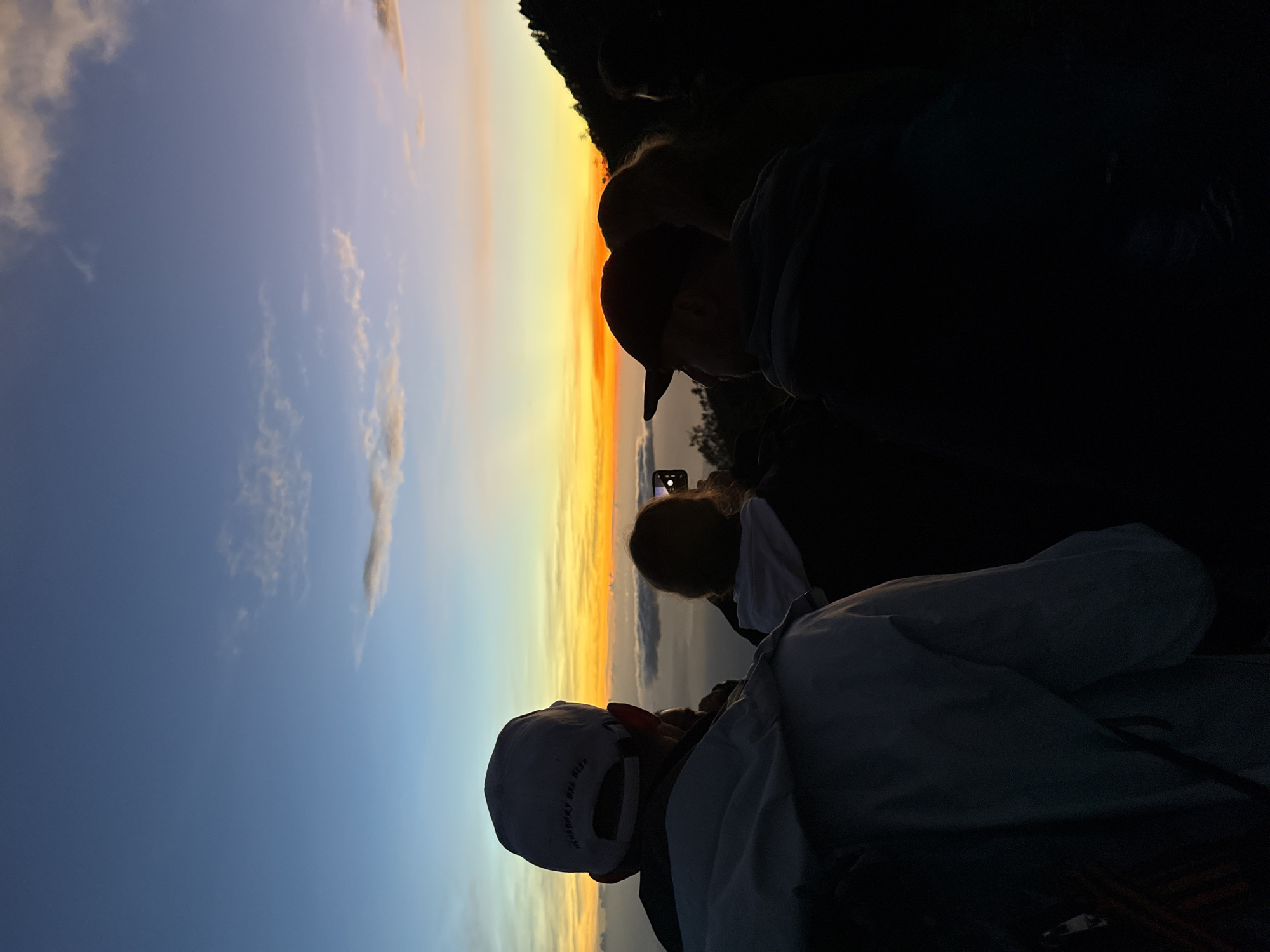 At an unknown location in Indonesia, a group of people are watching a sunset. The sky transitions from blue to orange. The people are mostly silhouetted. One person is holding a phone and taking a picture of the sunset. A person in the foreground is wearing a white baseball cap.