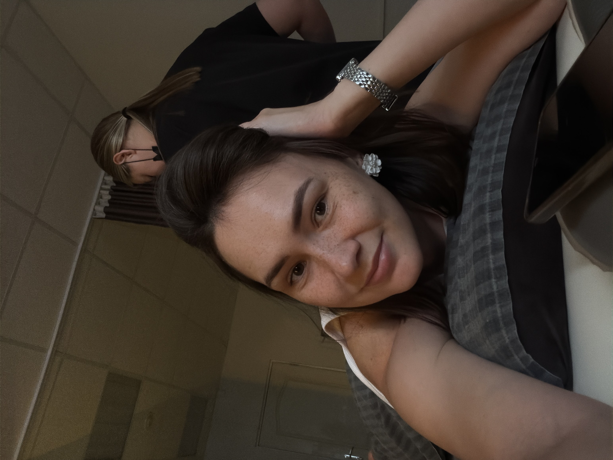 The image is a selfie taken by a young woman with dark hair and freckles. She is lying on her side on a pillow and smiling at the camera. Her left hand is resting on her head, and she is wearing a silver watch on her right wrist and a flower-shaped earring in her left ear. A second person, with blonde hair pulled back in a ponytail and wearing a black shirt and a black face mask, is visible in the background, turned away from the camera. The environment appears to be an indoor setting with a tiled ceiling and a door in the background. The scene does not provide any specific details that would link it to the city of Chaikovsky, Russia.
