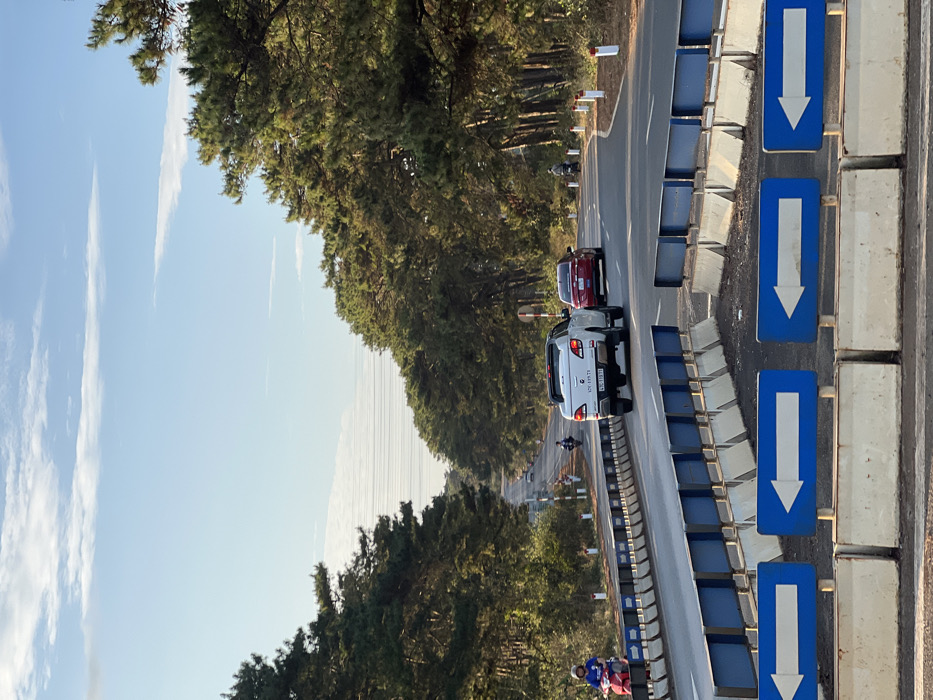 The image shows a multi-lane asphalt road traversing a dense evergreen forest, located in Ea Ksô, Vietnam, under a bright blue sky with scattered white clouds. In the immediate foreground, a line of light-colored concrete barriers topped with blue panels features four blue rectangular signs, each displaying a white left-pointing arrow. The road curves gently to the right as it extends into the distance, with continuous concrete barriers and blue panels lining its left side and small white and red reflector posts marking the right edge. A white pickup truck, bearing license plate 47C-338.79, is positioned in the right lane, moving away from the viewer. A red sedan follows behind it in the same lane. Three individuals on motorcycles are visible: one person on a red scooter is on the far left shoulder, adjacent to the barriers; another person on a dark motorcycle is further down the road near the center; and a third individual on a dark motorcycle is on the far right shoulder. Dense evergreen trees, characteristic of a pine forest, line both sides of the road. Faint power lines are visible spanning across the distant sky, above the road, between the tree lines.