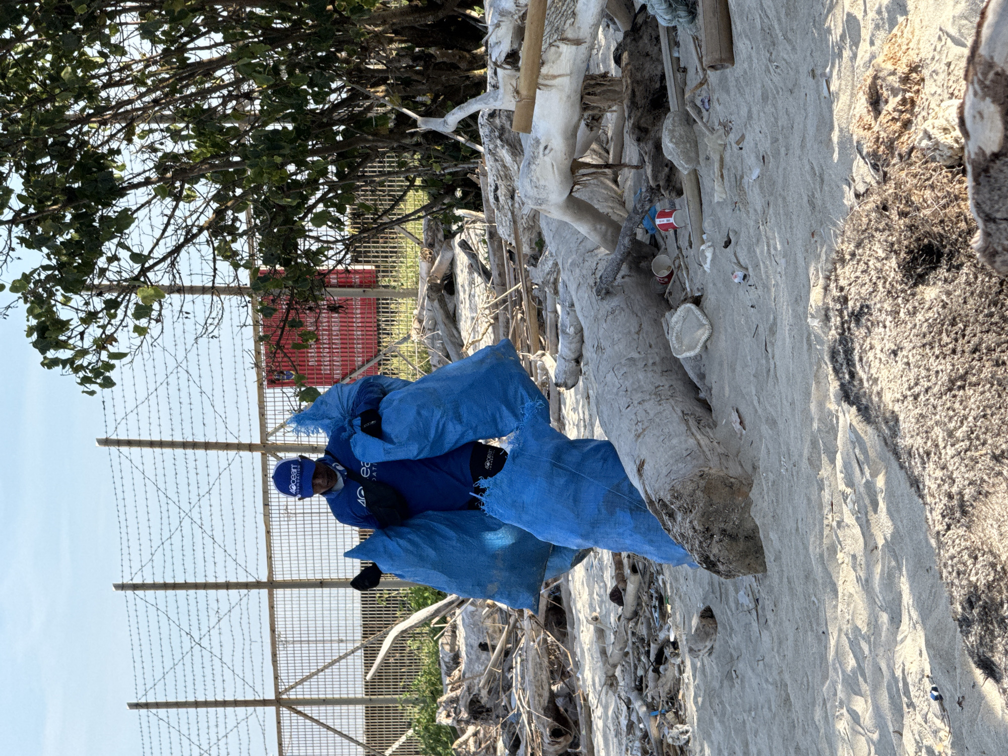 A person wearing a blue hat and shirt with the "4ocean Foundation" logo is carrying three blue bags filled with trash on a sandy beach. The beach is littered with trash, driftwood, and other debris. A wire fence with trees and a red building are visible in the background. The location appears to be a polluted section of a beach in Denpasar, Indonesia.