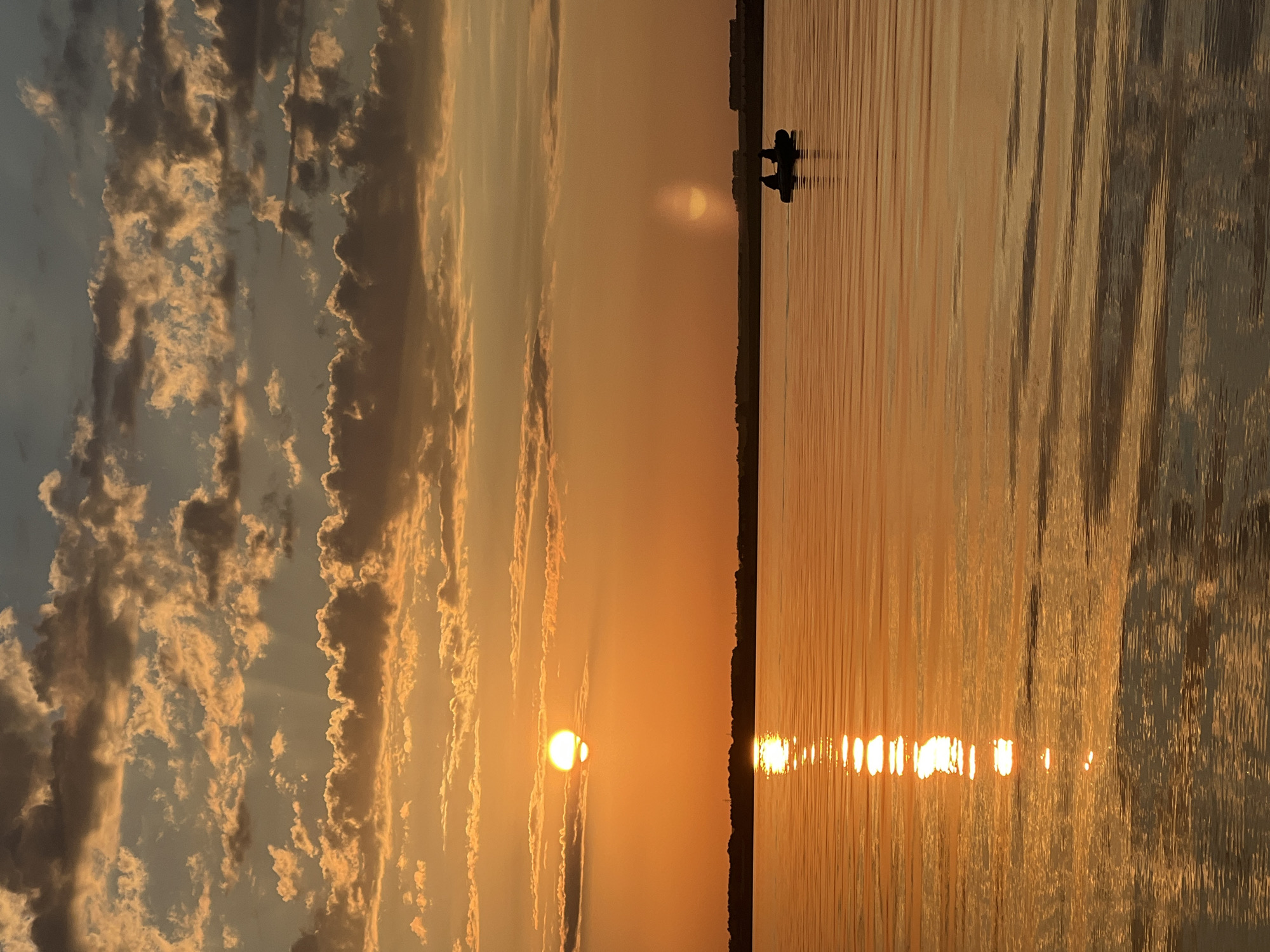 A sunset over a body of water, possibly a lake or wide river, is depicted. The sky is filled with orange and golden clouds, with the sun a bright, distinct circle peeking through. The water reflects the warm colors of the sky, creating a shimmering effect. In the distance, a dark silhouette of land is visible, likely a treeline. Closer to the foreground, a small, dark inflatable boat with two silhouetted figures is on the water. The scene appears peaceful and serene, with the golden hour light casting a warm glow over the landscape. The location is identified as Novopichugovo, Russia.