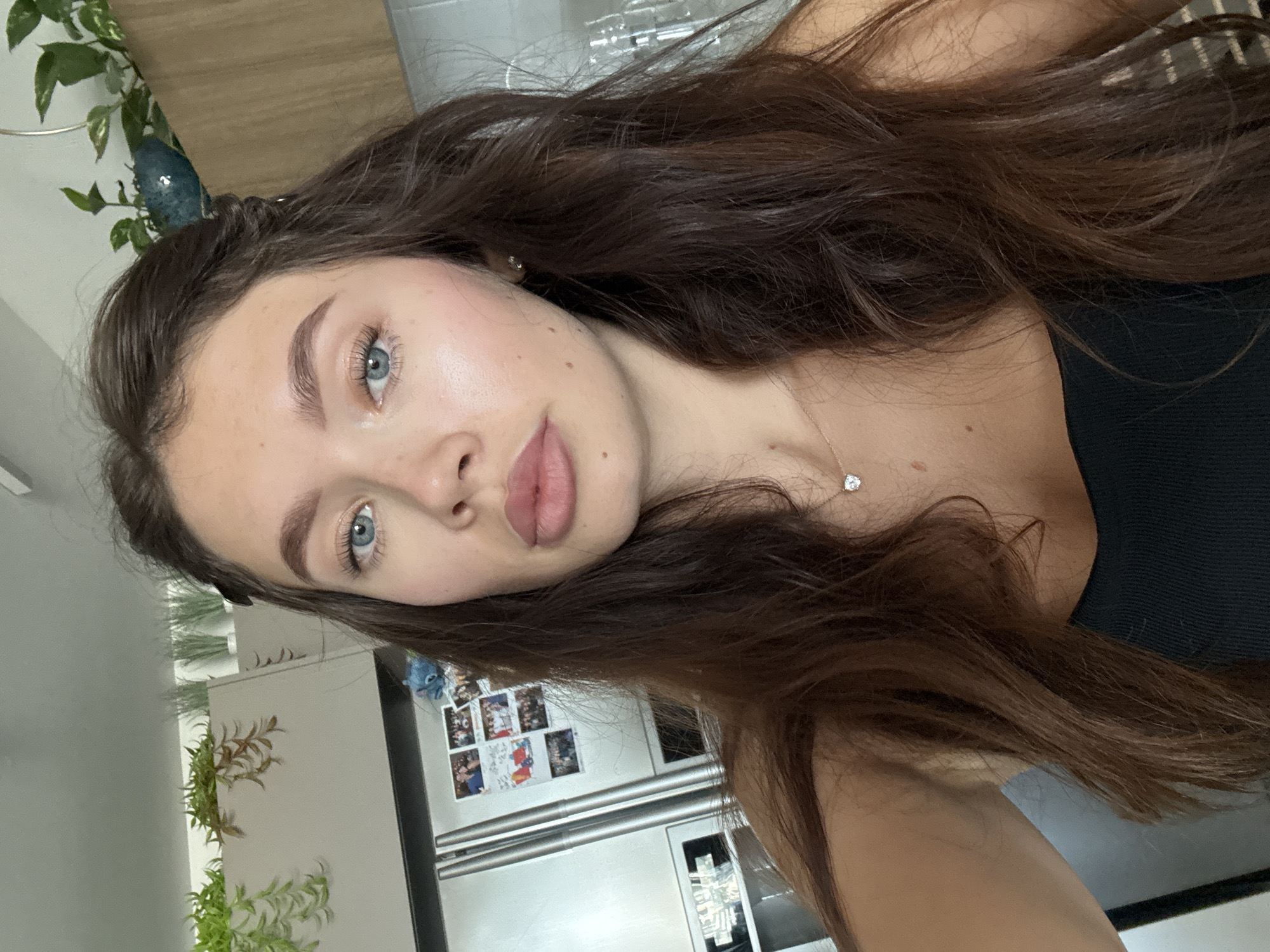 A young woman with long brown hair, blue eyes, and light skin is centered in the frame, appearing to take a selfie. She is wearing a black top and a delicate necklace with a small pendant. The background shows a kitchen interior with white cabinets, a stainless steel refrigerator covered with photographs and magnets, and various potted plants. The location is a residential kitchen, inferred, but there is no location of it in Tel-Aviv.