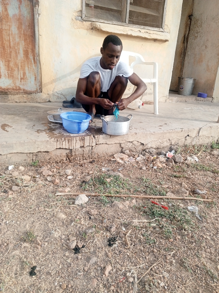 A young, dark-skinned man is depicted squatting outdoors in Kuta, Nigeria, engaged in a washing activity. He wears a white t-shirt and dark shorts, his gaze fixed on a blue-green piece of material he is handling inside a silver metal pot. A blue plastic bowl, also containing water, sits beside the pot, and spilled water is visible on the concrete slab where he is positioned. A pair of dark sandals are behind him on the ground.
The scene unfolds in front of a weathered building with a light-colored wall. To the left, a rustic wooden door is visible, and above the man, a window with a metal frame can be seen. On the right side of the frame, a white plastic chair stands empty, and further back, a silver metal bucket with the Chinese characters "新发 不锈" (Xinfa Stainless) and a white plastic jug are present.
The foreground consists of dry, unpaved ground littered with small rocks, patches of sparse dry grass, and various debris including sticks. The overall lighting indicates daytime, likely late morning or afternoon, with clear weather conditions. The man appears focused and composed while performing his task in this practical, outdoor setting.