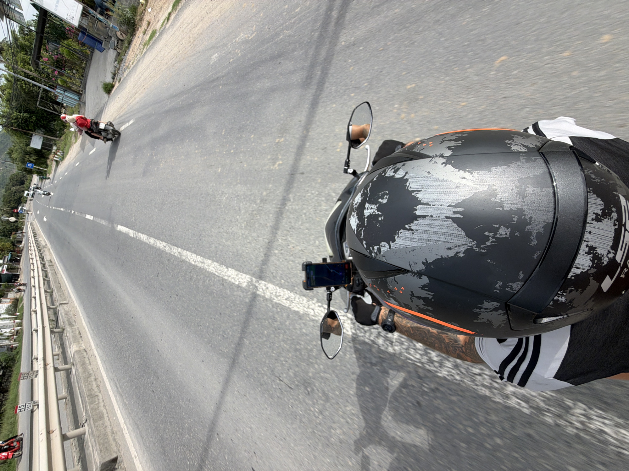 The image provides a first-person perspective from a motorcycle rider, looking forward down a paved road on a bright, sunny day. The scene is dominated by the asphalt road, stretching into the distance under clear skies, flanked by greenery and some roadside structures.
In the foreground, the top of a black motorcycle helmet, worn by the person taking the photograph, is prominent. The helmet features a distinctive silver/gray abstract, distressed pattern over a matte black base, with a subtle orange accent stripe visible towards the back. Partially visible text on the helmet's side suggests "LS2". The rider's right arm, adorned with tattoos and wearing a black glove and a smartwatch, is visible near the handlebars. A smartphone, displaying what appears to be a navigation application, is mounted on the handlebars. Rearview mirrors reflect parts of the road and the rider's arm. The rider is wearing a white t-shirt with black stripes.
Ahead on the road, another person on a red scooter or motorcycle is visible in the middle distance, also traveling in the same direction. The road itself is wide, with clear white dividing lines. To the left, a metal guardrail separates the road from a lower, parallel service road or shoulder, and beyond that, lush trees and some buildings line the route. Roadside signs with numbers "55" and "88", likely indicating speed limits or distance markers, are visible on the left side of the road. The background features more distant hills or mountains covered in vegetation. The setting, characterized by the visible road signs and environmental features, aligns with the provided context of Nam Cam Ranh, Vietnam.