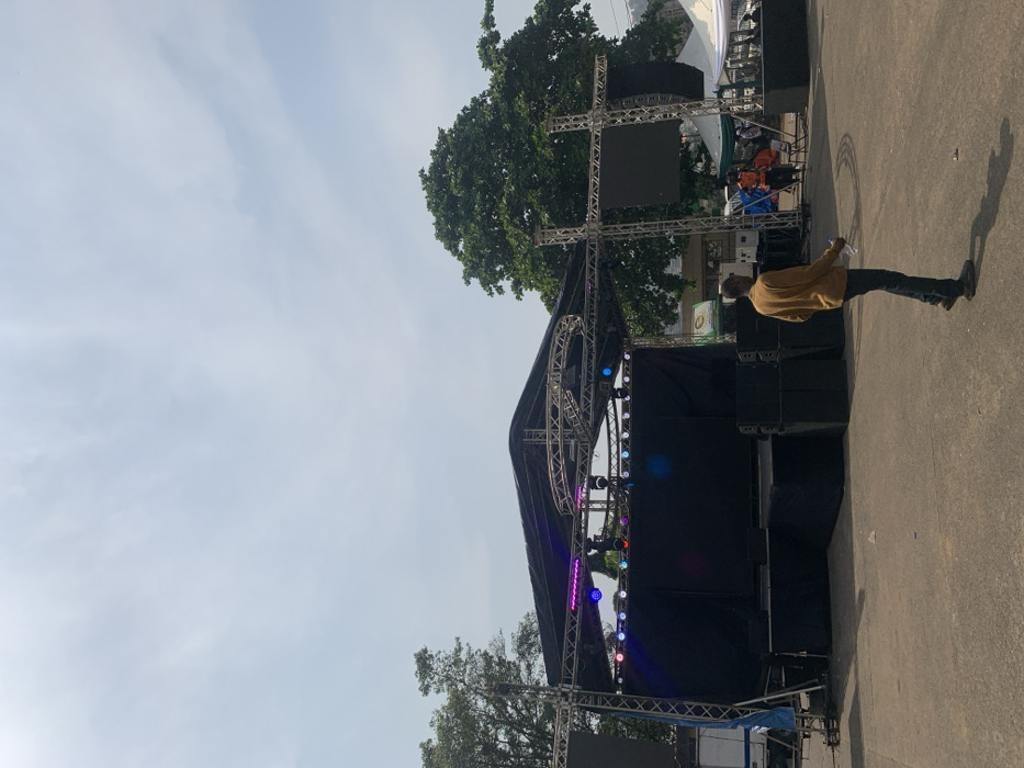 The image, oriented correctly, displays an outdoor event setup under a clear, bright daytime sky, likely in Freetown, Sierra Leone. A large, complex stage structure dominates the scene, constructed from metal trussing and covered in black fabric. Numerous speaker stacks are positioned on and around the stage, and a variety of colorful lighting fixtures, including LED par cans and moving heads, are mounted on the trussing, emitting blue, pink, and purple lights. A large, dark screen is visible on the upper left side of the stage structure.
In the immediate foreground, a man wearing a mustard yellow long-sleeved shirt and dark trousers stands with his back to the viewer, facing the stage. He holds a clear plastic bottle in his left hand, and his shadow is cast to his right on the paved ground. Lush green trees are visible behind and to the left of the stage, and another white tent structure, along with a few indistinct figures, can be seen in the background. A small white truck is partially visible near the lower left of the stage. A small, blurry green and white banner with unreadable text is in the far background.
The scene suggests the setup or a quiet moment before a public event, such as a concert, festival, or gathering, with the stage, sound system, and lighting prepared for use.