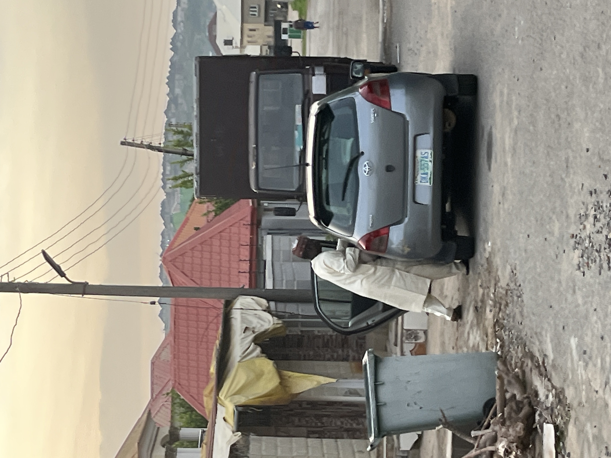 In Abuja, Nigeria, a man in traditional white Islamic attire is seen leaning into the open driver's side door of a silver Toyota Yaris. The car's license plate reads "DKA 557AS." Behind the Yaris, a dark-colored truck is parked, its front partially obscured. To the left of the scene, a large, light-grey garbage bin sits next to a structure covered by a yellow tarp. The environment suggests a residential or commercial street with buildings and red-tiled roofs visible in the background. Power lines and a street lamp are present overhead. The ground is paved with asphalt and debris is scattered in some areas. In the distance, a person in blue attire walks away from the viewer. The sky is overcast with a pale yellow hue.