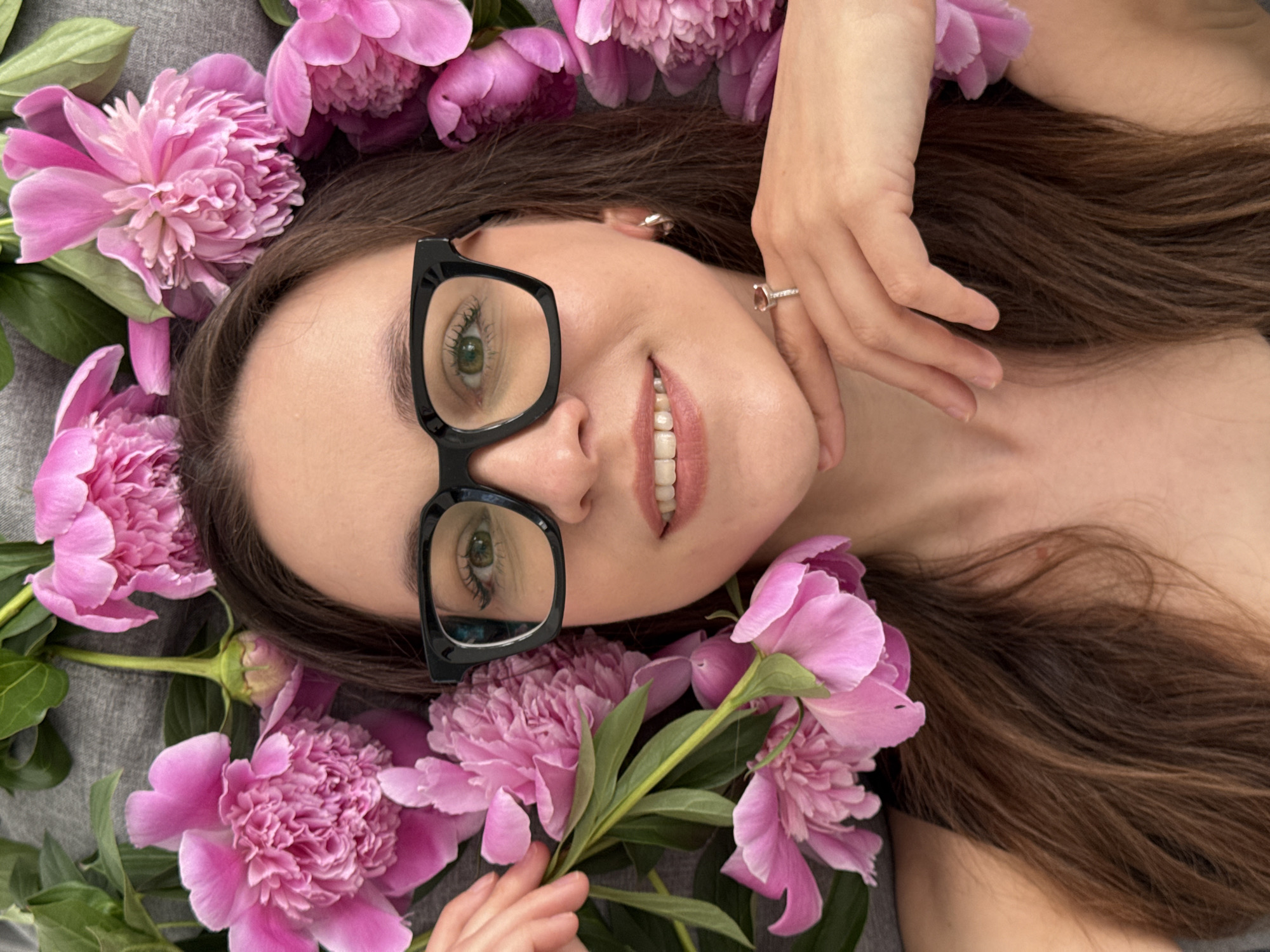 A woman with light skin, brown hair, and black-framed glasses is lying down, surrounded by pink peonies. She is smiling, with her right hand near her chin, showing a ring. The background is a gray fabric surface. The image contains no identifiable information about the city of Chaikovsky, Russia.