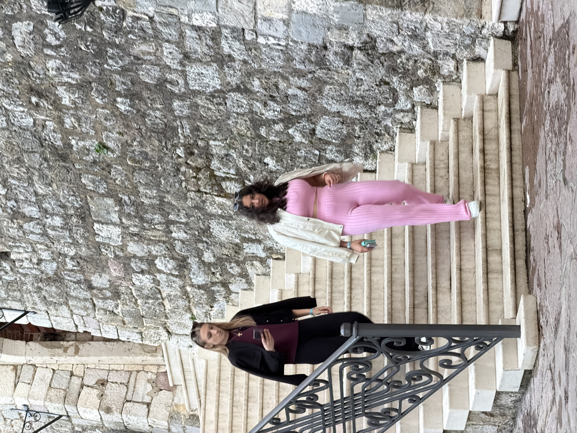Two women pose on a stone staircase in Budva, Montenegro. The staircase is constructed with light-colored stone steps and bordered by a rugged stone wall on one side and a stone wall with a metal railing on the other. One woman, positioned at the bottom of the frame, is dressed in a maroon top and black pants with a black blazer. She holds a mobile phone. The other woman, higher up on the staircase, wears a pink matching set, a white jacket, and sunglasses on her head. The background consists of the stone walls and stairs.