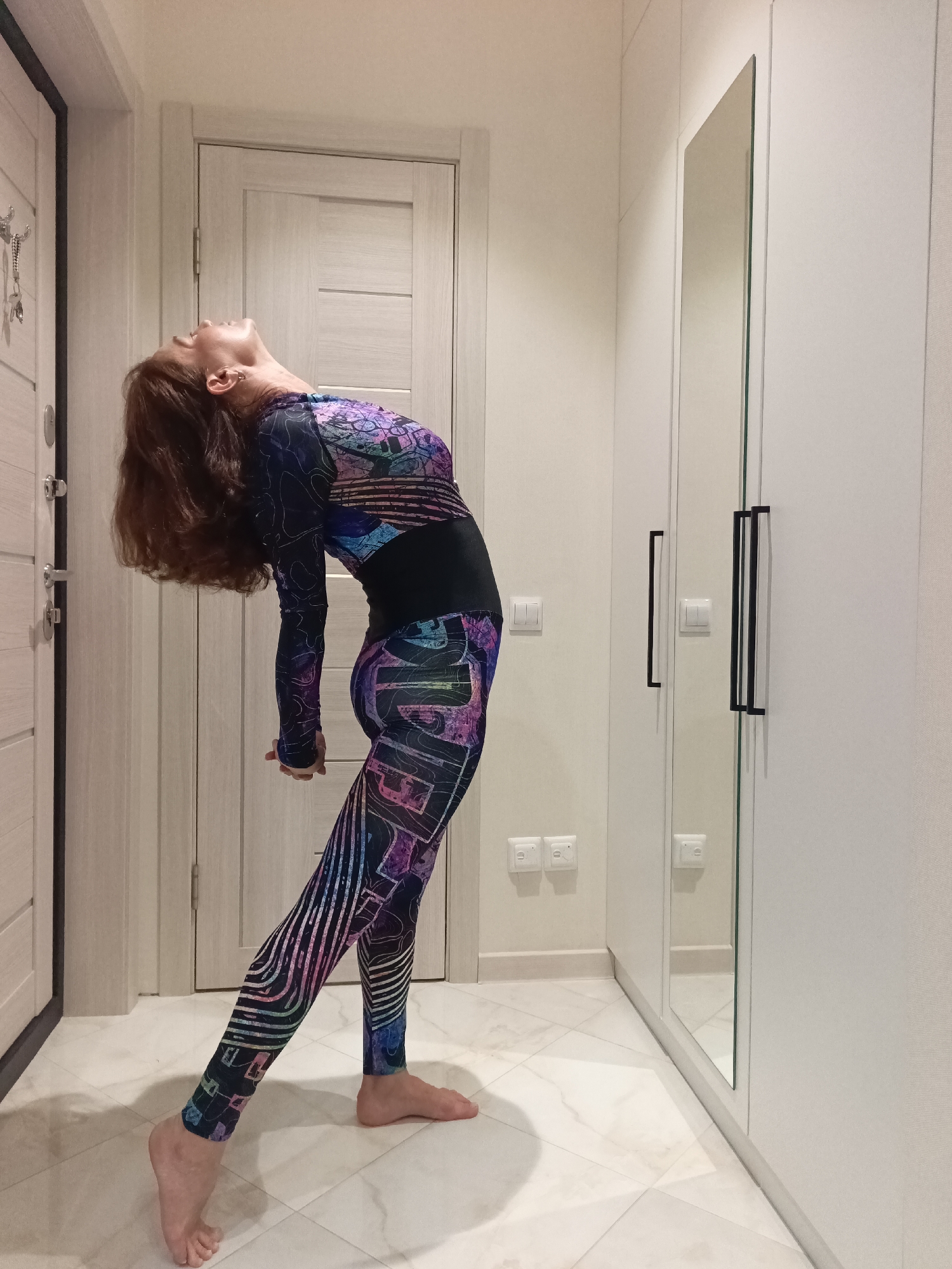 A woman with long brown hair is performing a backbend in a hallway. She is wearing a full-body activewear suit with a colorful geometric and abstract pattern. Her left leg is extended forward and slightly bent, while her right leg is extended backward, with her right foot raised on the ball of her foot. Her arms are clasped behind her back, and her head is tilted back. The hallway has white walls and doors, a light-colored tiled floor with a marble pattern, and a tall white wardrobe with mirrored doors on the right side. Light switches and electrical outlets are visible on the walls. The scene appears to be indoors, likely in a residential setting. There is no indication that this image was taken in Sochi, Russia.