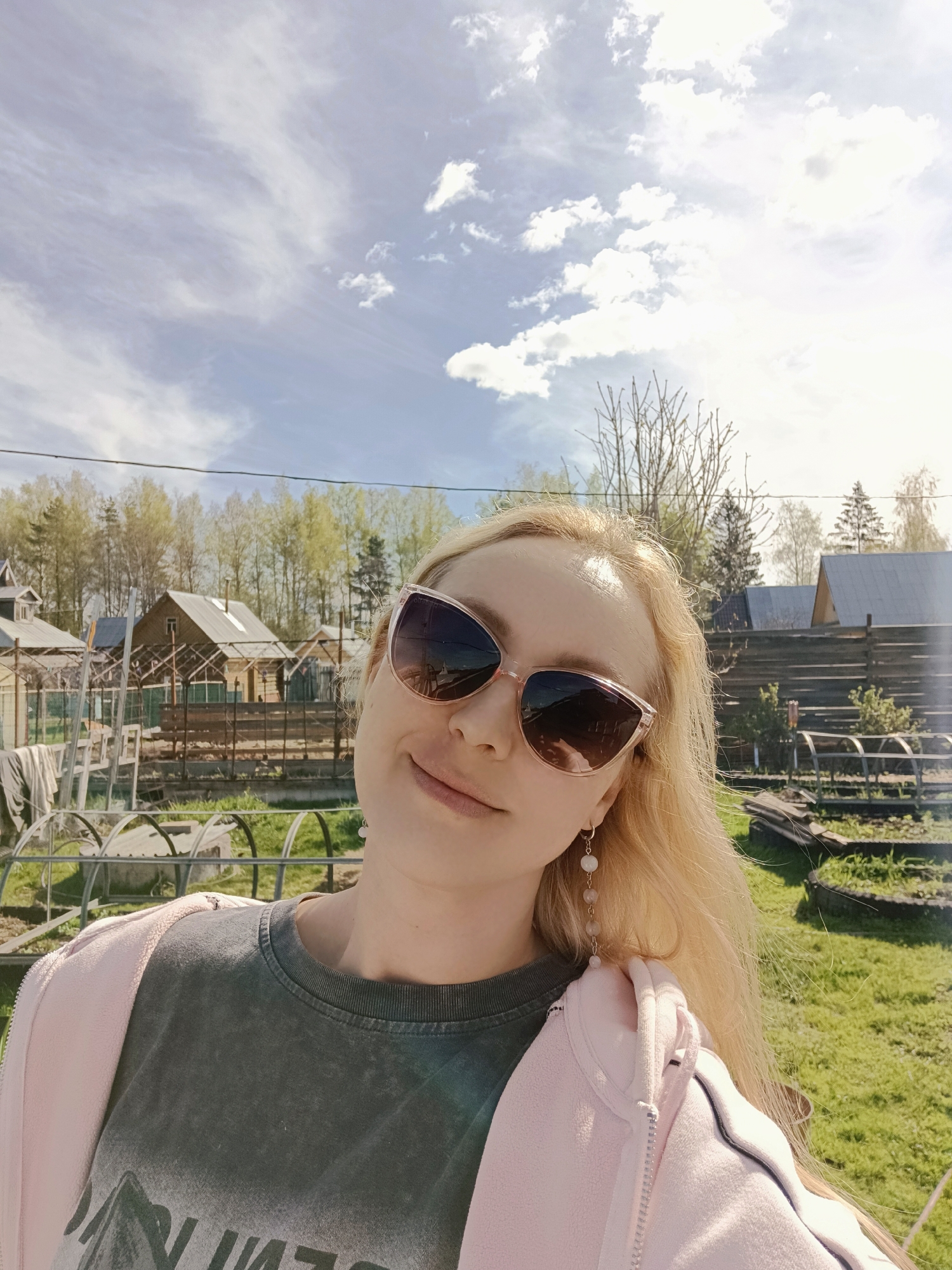 A selfie of a blonde woman wearing sunglasses, a gray t-shirt, and a pink jacket, taken outdoors in Вичелово, Russia. In the background are houses, trees, a garden area with arched frames, and a sky with scattered clouds.