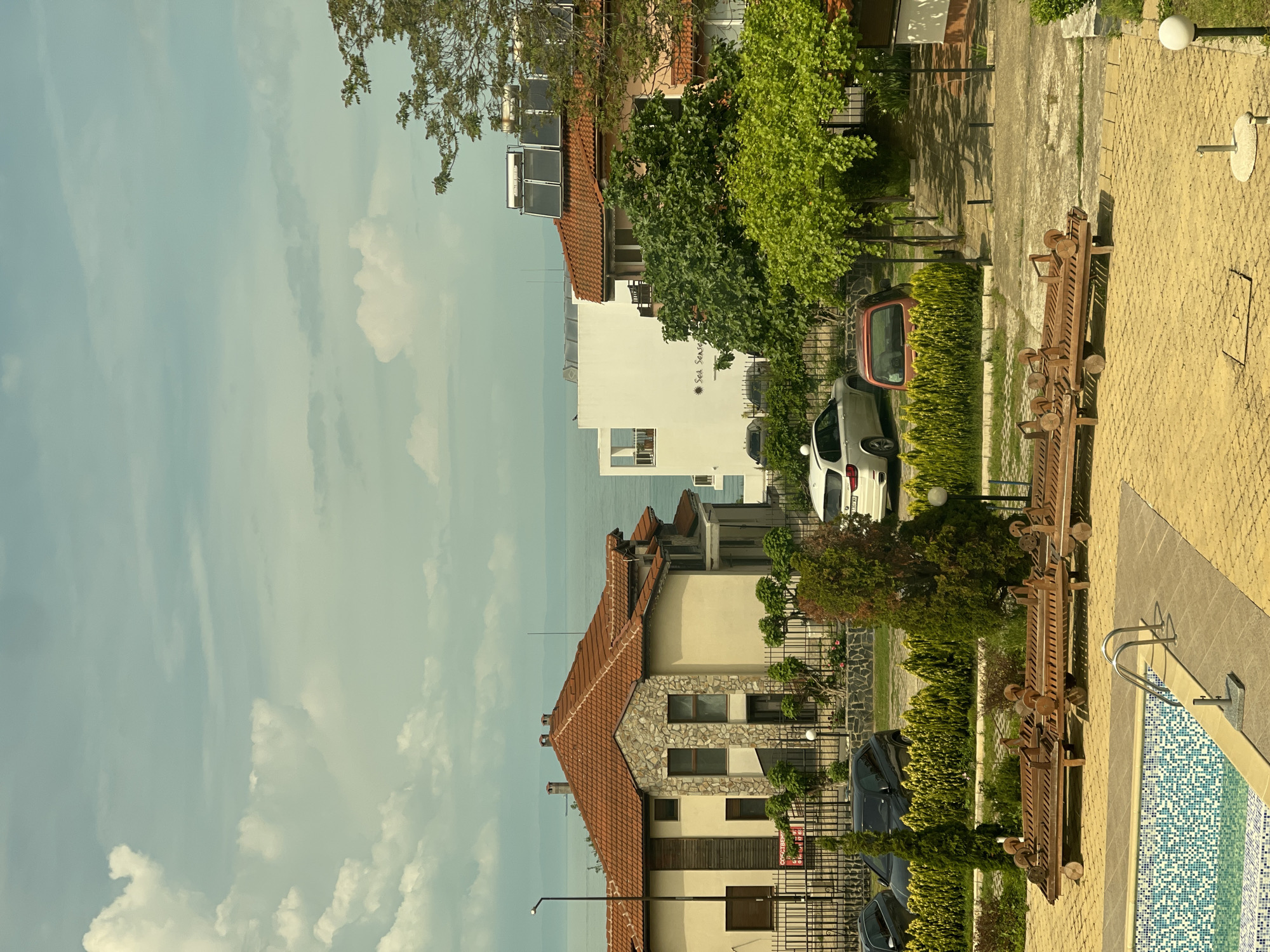 The image depicts an outdoor scene in Sozopol, Bulgaria, featuring buildings, vehicles, and landscaping. The sky is visible with a light blue hue and scattered clouds. A multi-story building with a red-tiled roof and a stone facade is prominent in the foreground. Adjacent to it is a white building. Several vehicles, including cars, are parked along a driveway or street. Lush greenery and landscaping elements such as bushes and trees are present. A pool is visible to the right. Architectural details include fences, solar panels on rooftops, and exterior lighting. The overall impression is of a residential or resort area in a coastal town.