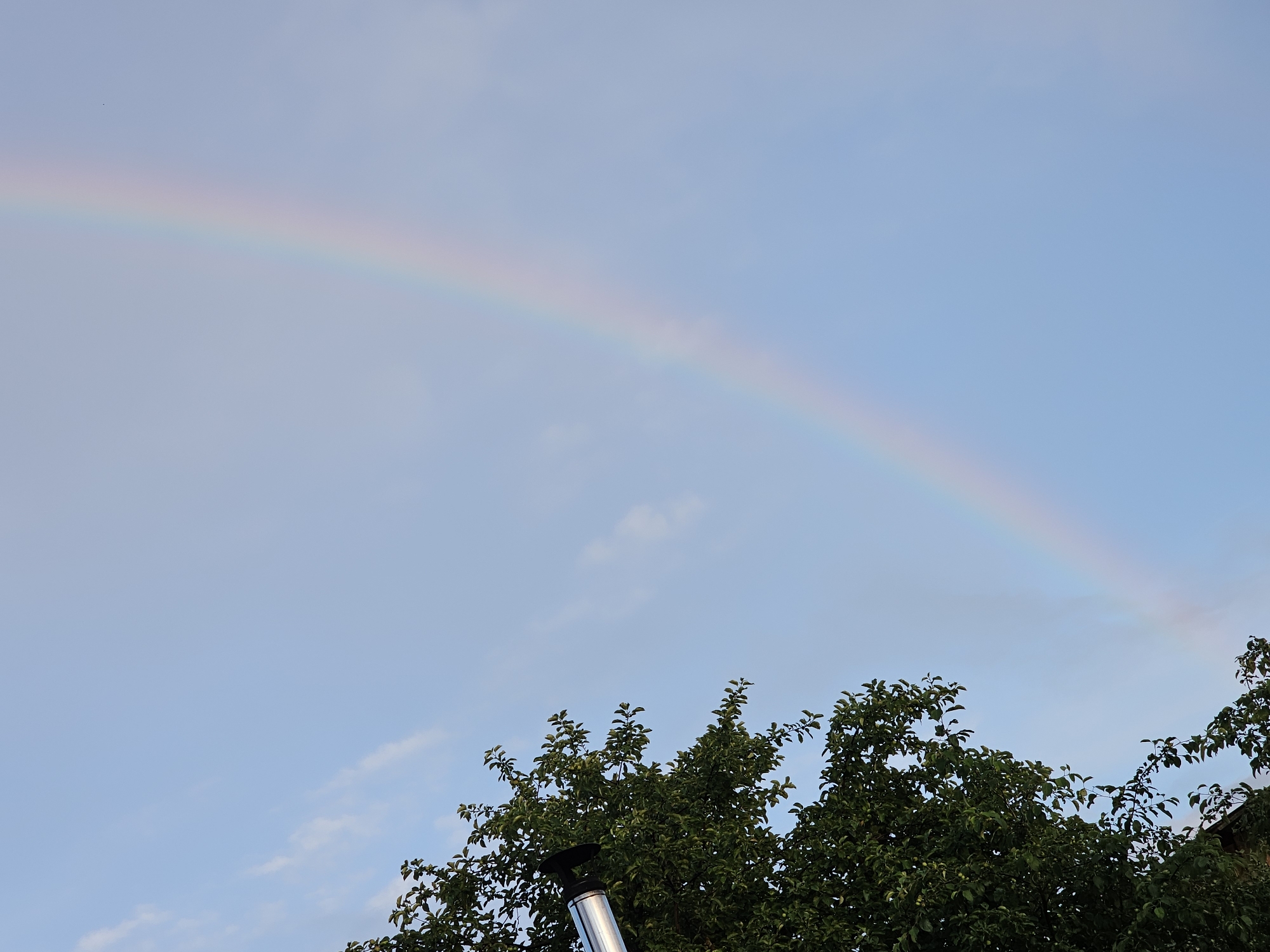 The image captures a clear sky with a faint rainbow arching across it. The lower portion of the frame is dominated by the dense green foliage of trees, suggesting an outdoor scene. A metal chimney pipe protrudes from the bottom left of the image, partially obscured by the leaves. The location is identified as Альняш, Russia.