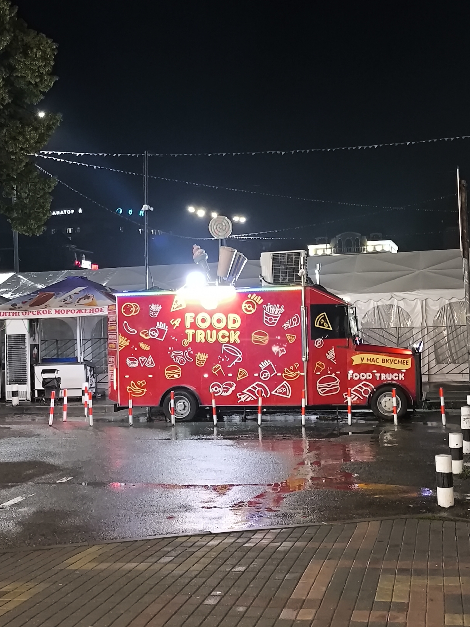 A red food truck, adorned with cartoon images of food and the words "FOOD TRUCK," is parked on a wet street at night. The food truck has a large advertisement on its side that reads "У НАС ВКУСНЕЕ FOOD TRUCK," which translates to "We Have Tastier FOOD TRUCK." The street is illuminated by the lights of the food truck and string lights overhead, which are decorated with small triangular flags. In the background, there is a white tent structure and a building with illuminated windows. To the left of the food truck is a smaller kiosk with a blue awning that has text on it, and what appears to be a small white freezer unit. The ground is paved with cobblestones and asphalt, both of which are wet, indicating recent rain. Several orange and white striped bollards are placed around the food truck. The scene appears to be a public gathering or market area in Yessentuki, Russia, likely at an outdoor event or promenade.