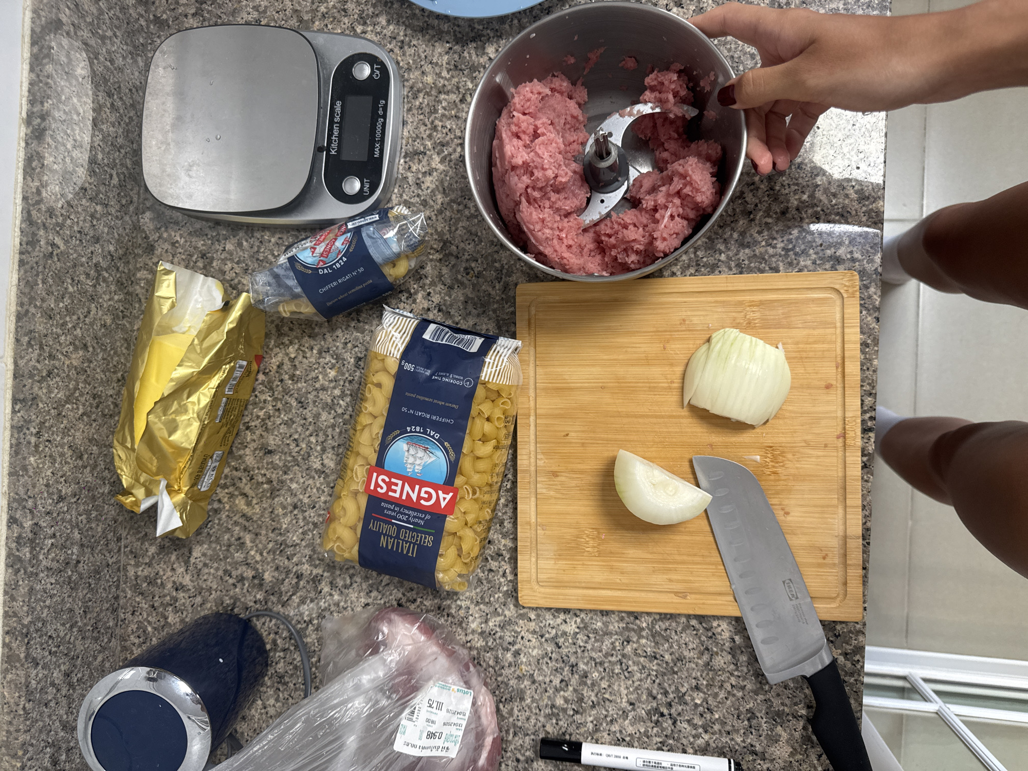 The image shows a kitchen counter with various food preparation items. A kitchen scale is on the upper left. Next to it are two opened packages of pasta, one gold and one blue, and the blue one is labeled "AGNESI CHIFFERI RIGATI N° 50". A stainless steel bowl filled with minced meat and a blade attachment is in the upper right corner, partially held by a hand. A wooden cutting board with a chopped onion and a knife lies on the counter. There's also a package of meat and a blender base at the bottom of the image, as well as a black marker. A person wearing socks is visible standing next to the counter. The presence of "Lotus's" branding on the meat package and the "AGNESI" pasta suggests a location in Chalong, Thailand, as Lotus's is a retail chain with a presence there.