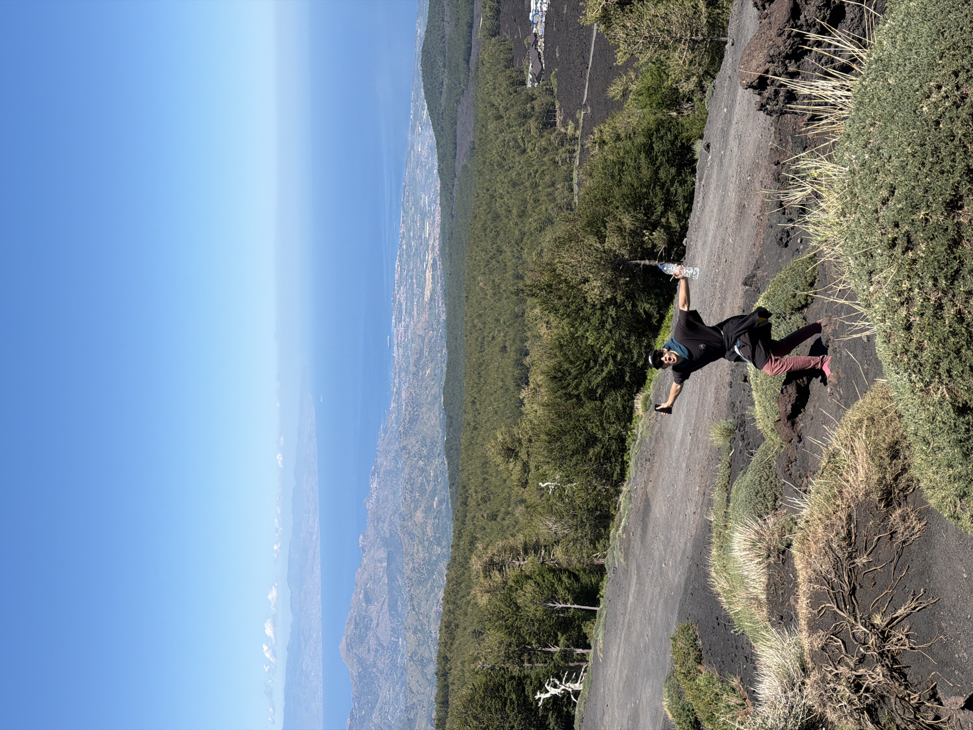 A person with a dark t-shirt, burgundy pants, and pink shoes stands on a dirt path on a mountainside, holding a plastic water bottle and a smartphone. The person is looking toward the camera with an open mouth. In the background, a vast green forest stretches out towards a hazy coastline and the blue ocean under a clear sky. The terrain is dark and rocky, with sparse vegetation in the foreground.
