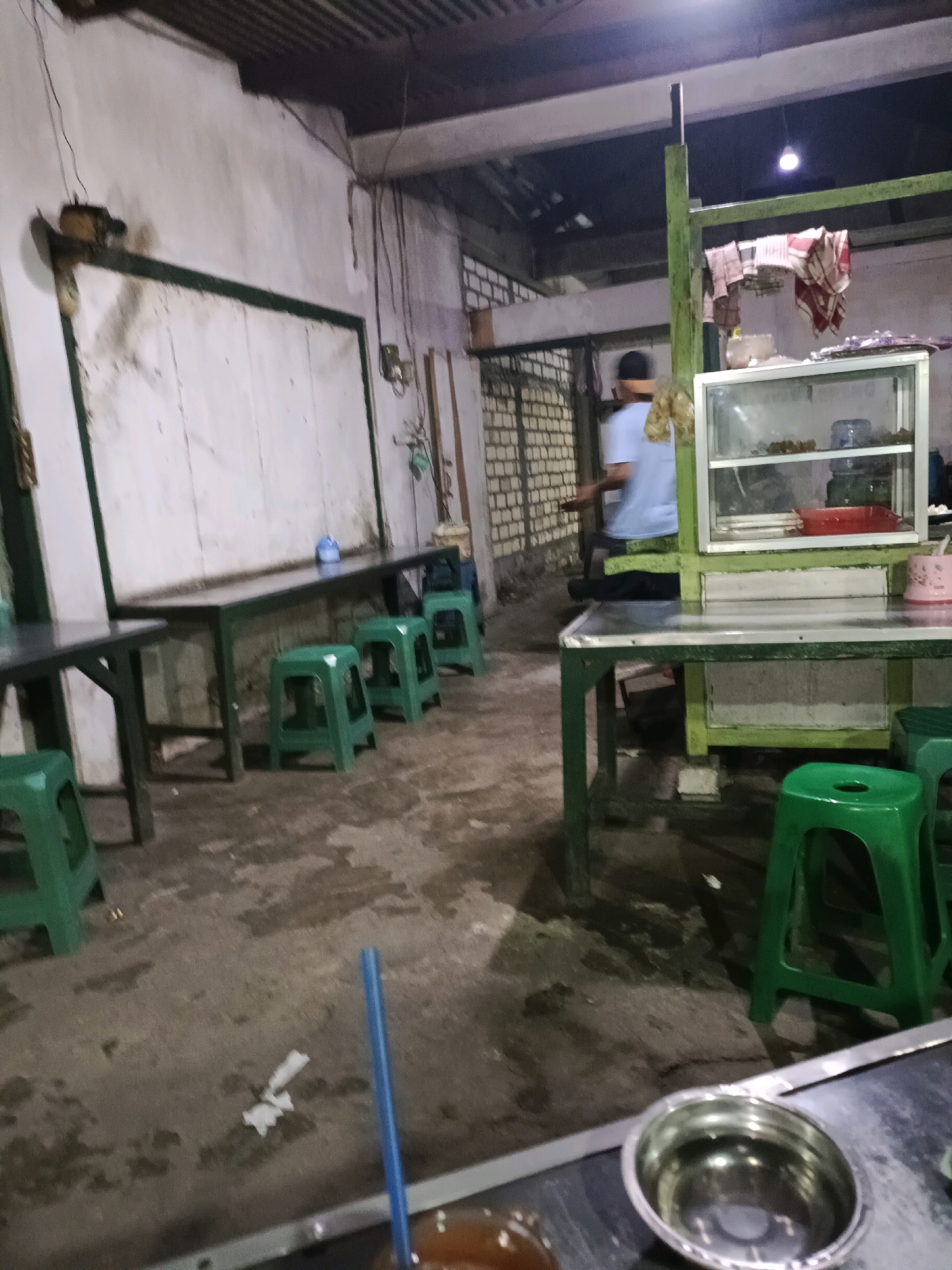The image depicts an indoor scene, likely a food stall or small eatery, within Sukomulyo, Indonesia. The setting appears utilitarian and somewhat rustic. A long, dark table with a green metal frame runs along the left side of the frame, with several green plastic stools arranged in front of it. A similar setup is visible on the right side of the image, with a green metal frame supporting a display case containing food items and a red plastic container.
A person, wearing a light blue shirt and a dark cap, is visible in the background, facing away from the camera. They appear to be working or serving customers. The walls are a light color, possibly painted concrete or plaster, with some visible brickwork in the background. The floor is made of concrete and appears wet or stained in places. Overhead, a corrugated metal roof is visible, with hanging wires and a light fixture. A blue drinking straw is prominently featured in the foreground, along with a metal bowl. The overall impression is of a busy, informal dining establishment.