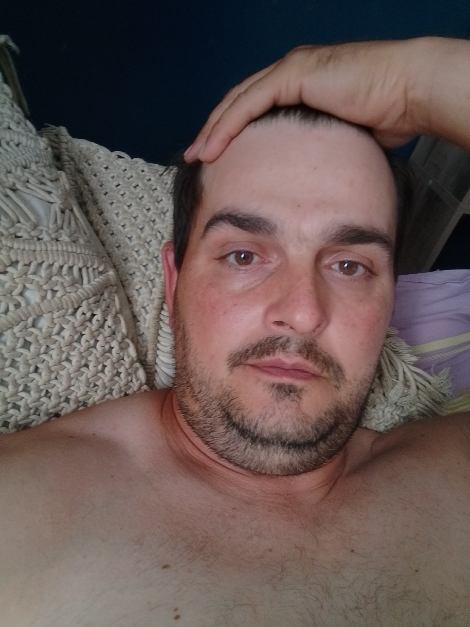 A man with fair skin, a light beard, and brown eyes is lying down with his hand on his head. He is shirtless and looking at the camera. He is in a bedroom, lying on a bed with white and purple bedding. A woven blanket is behind him. The scene is lit by natural light.