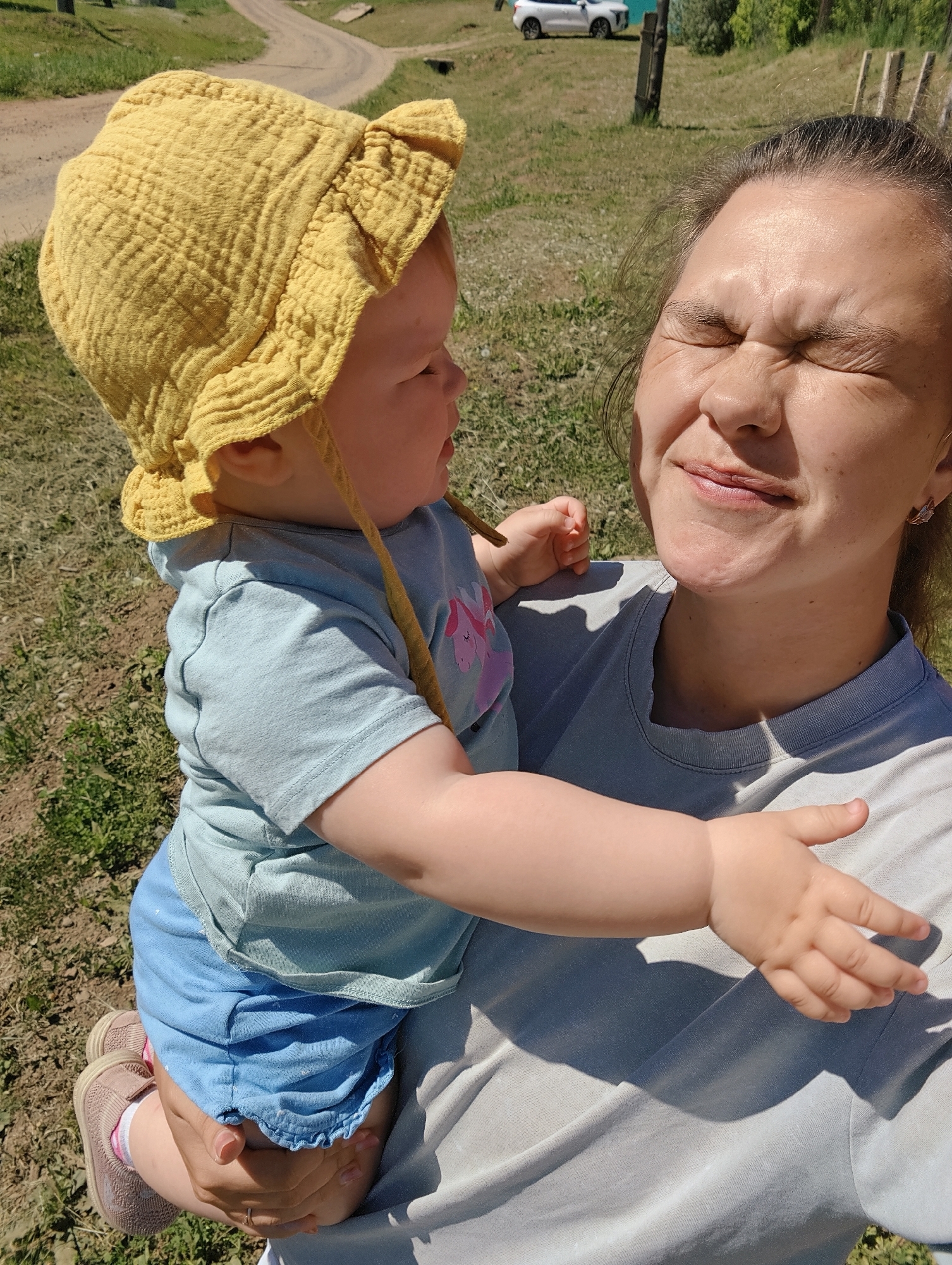 The image shows a fair-skinned woman holding a baby. The woman is squinting her eyes shut as if from bright sunlight and is smiling slightly. The baby, wearing a yellow bonnet, light blue shirt, and blue shorts, looks to the side. The background consists of grassy terrain and a dirt road leading away from the camera. A white vehicle is parked in the distance. The location is Большой Букор, Russia.