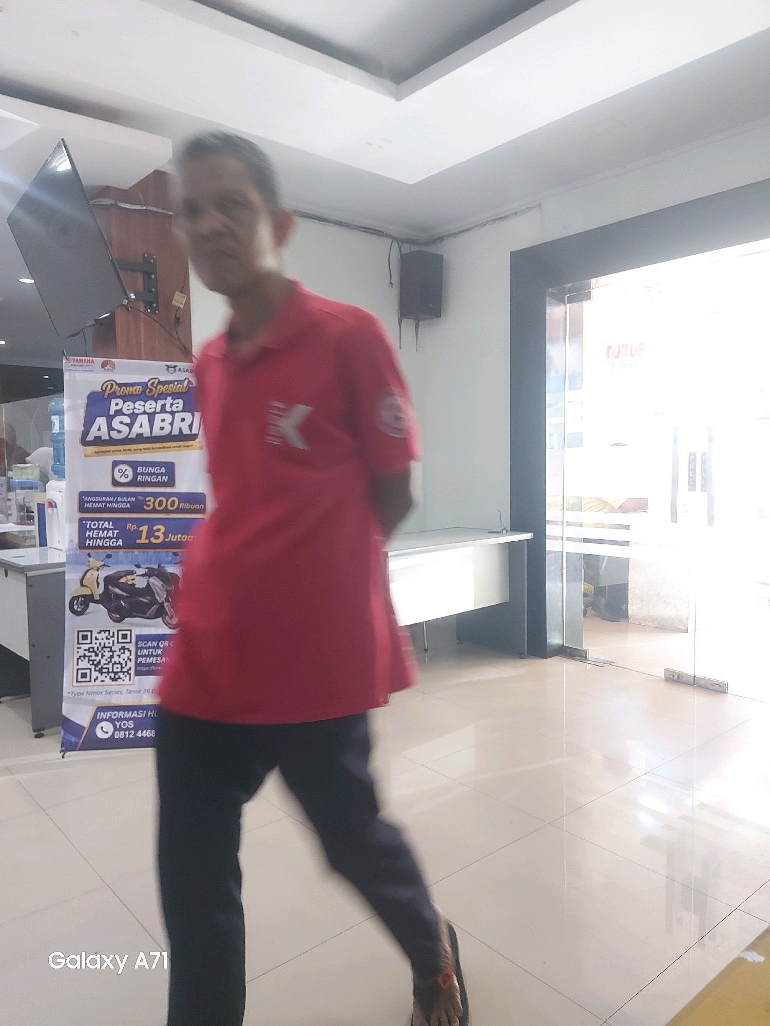 The image depicts a slightly blurred scene inside a bright, modern-looking commercial space, likely a showroom or office, with large windows and reflective tiled flooring. A man, the primary subject, is in motion, walking towards the right side of the frame. He is wearing a red polo shirt and dark trousers. To the left of the man, a large promotional banner is visible, featuring text in Indonesian. The banner displays information about a special promotion for "ASABRI" participants, detailing installment plans for motorcycles, with images of two scooters. A water cooler is also present behind the banner. On the right side of the image, a glass door entrance is visible, with "TUTUP" (Closed) text in red lettering on the glass. The overall lighting is bright, suggesting daytime. The presence of the "Galaxy A71" watermark in the bottom left corner indicates the photo was taken with a Samsung Galaxy A71 smartphone. While the exact location in Makassar, Indonesia, cannot be determined from the image alone, the Indonesian language on the banner and the general aesthetics suggest a commercial establishment within the city.