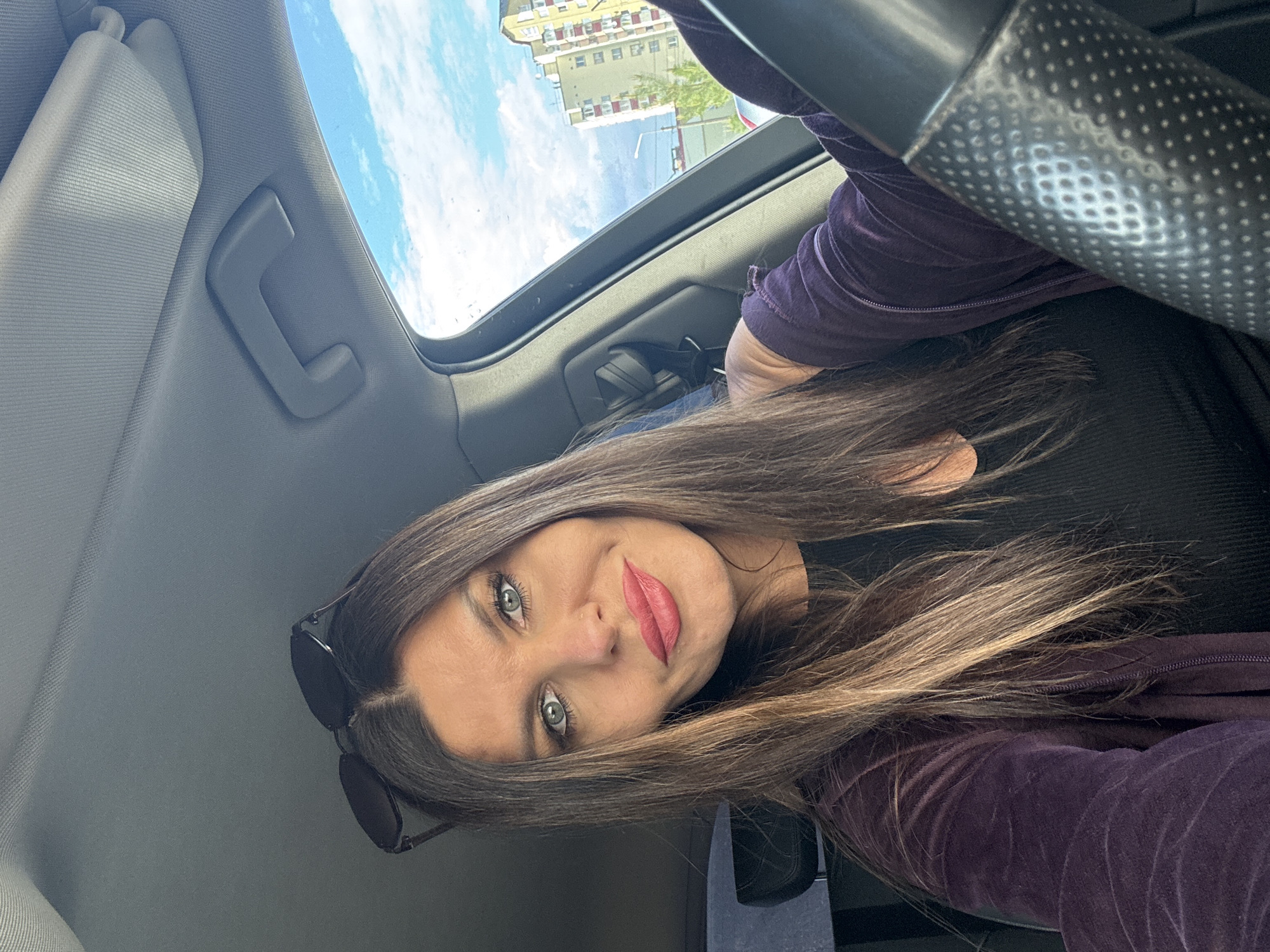 A woman is captured in a selfie from the chest up, seated in the driver's seat of a car. She has long, dark brown hair with blonde highlights and striking blue eyes. Her lips are painted a vibrant pink. She wears a dark purple velvet jacket over a black top. Sunglasses are perched on top of her head. The interior of the car is visible, including a portion of the steering wheel and the passenger-side interior. Through the car window, a bright blue sky with white clouds is seen, along with a yellow apartment building with red-trimmed windows, characteristic of Soviet-era architecture. This setting suggests the photo was taken in Chaikovsky, Russia, given the architectural style and cloud formations.