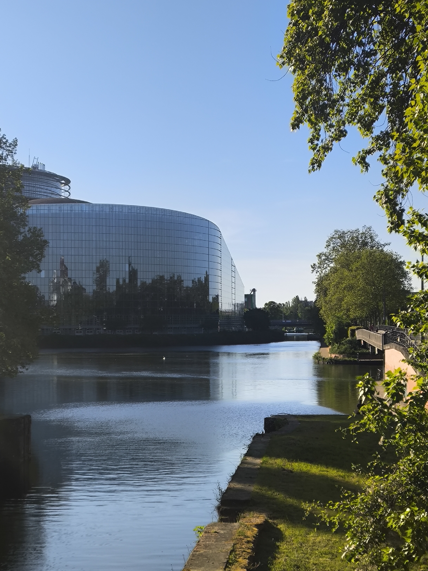 The image captures the Louise Weiss building, a part of the European Parliament complex located in Strasbourg, France. The building's distinctive, modern design features a large, curved glass facade that reflects the surrounding environment, including trees and other buildings. The building is situated along the Ill river, a waterway that adds to the scenery. The river is calm, with reflections of the building and trees visible on the water's surface. The foreground includes a grassy bank along the river's edge, with trees and greenery providing a natural frame to the scene. A bridge is also visible, adding to the architectural context of the location. The sky is clear and blue, indicating a sunny day. Overall, the image presents a tranquil urban landscape, highlighting the modern architecture of the European Parliament building within the natural and architectural setting of Strasbourg.