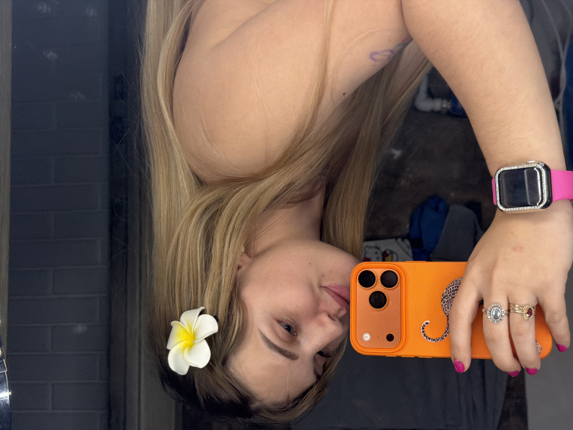 A woman is taking a selfie in a mirror. She has long blonde hair and is wearing a white flower in her hair. She is holding an orange iPhone with a leopard print sticker on it. She is wearing a pink Apple Watch and several rings. The background is a gray brick wall. The image appears to have been taken indoors, possibly in a bathroom. There is no clear indication of being in the city of Большая Шильна, Russia.