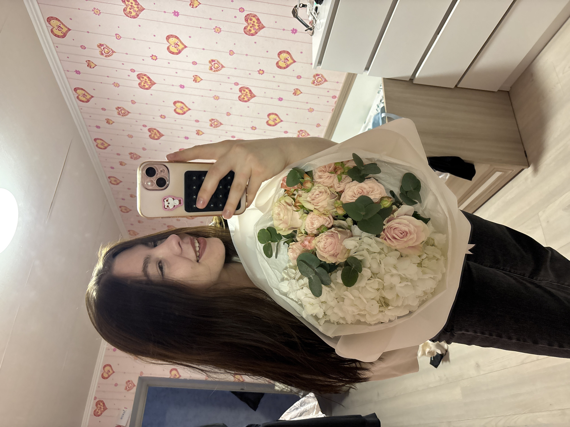 A young woman with long, dark brown hair is captured in a mirror selfie indoors, smiling broadly and showing her teeth. She holds a large, elegantly arranged bouquet of flowers in her left arm, featuring light pink roses, white hydrangeas, and green eucalyptus leaves, all wrapped in white paper. Her right hand holds a light-colored smartphone, possibly gold or rose gold, which has a distinct camera bump, a small white bear sticker, and a black grip accessory on its back. She appears to be wearing dark pants, possibly jeans.
The background reveals a room with a light pink wallpaper adorned with numerous pink and yellow heart patterns, interspersed with small pink floral or dot motifs. A white ceiling molding runs along the top edge of the wall. To the right of the woman, a white dresser or cabinet with multiple panels is visible, and below it, a light-colored wooden floor. A bright light source is partially visible on the far left, creating a reflection on the phone screen. The image conveys a happy and personal moment, likely celebrating the bouquet.