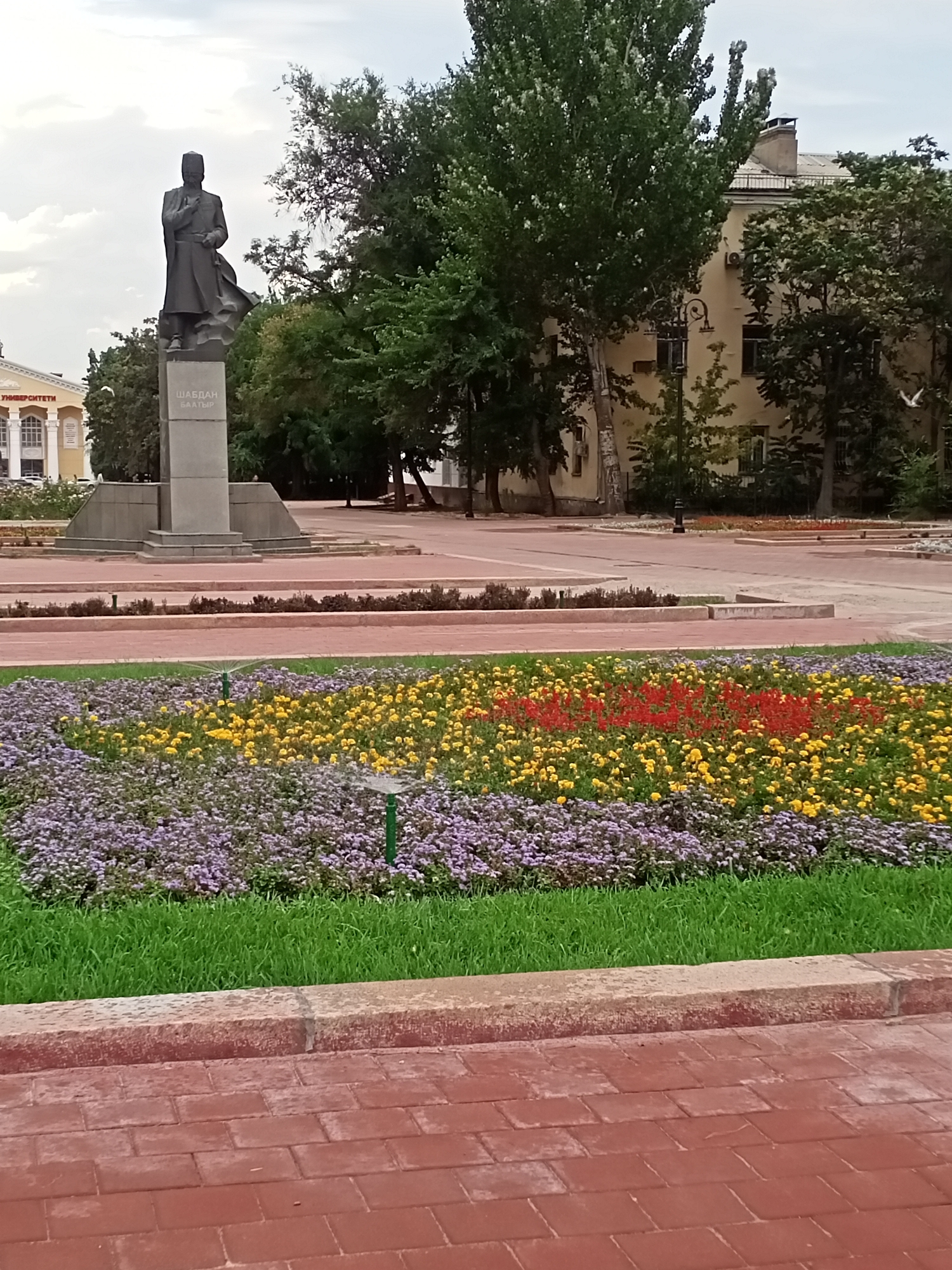 The image depicts a statue of a historical figure in a city square. The statue, made of dark metal, stands on a grey stone pedestal inscribed with text in a foreign language, likely Kyrgyz, reading "ШАБДАН БАТЫР". The statue is positioned in front of a building with light-colored walls and windows, partially obscured by trees with lush green foliage. In the foreground, a colorful flowerbed bursts with purple, yellow, and red blooms, with a sprinkler system visible amongst the flowers. The ground is paved with red brickwork, suggesting an urban environment. The sky above is overcast with scattered clouds. The overall scene suggests a public space in Bishkek City, Kyrgyzstan, possibly a park or a monument area.