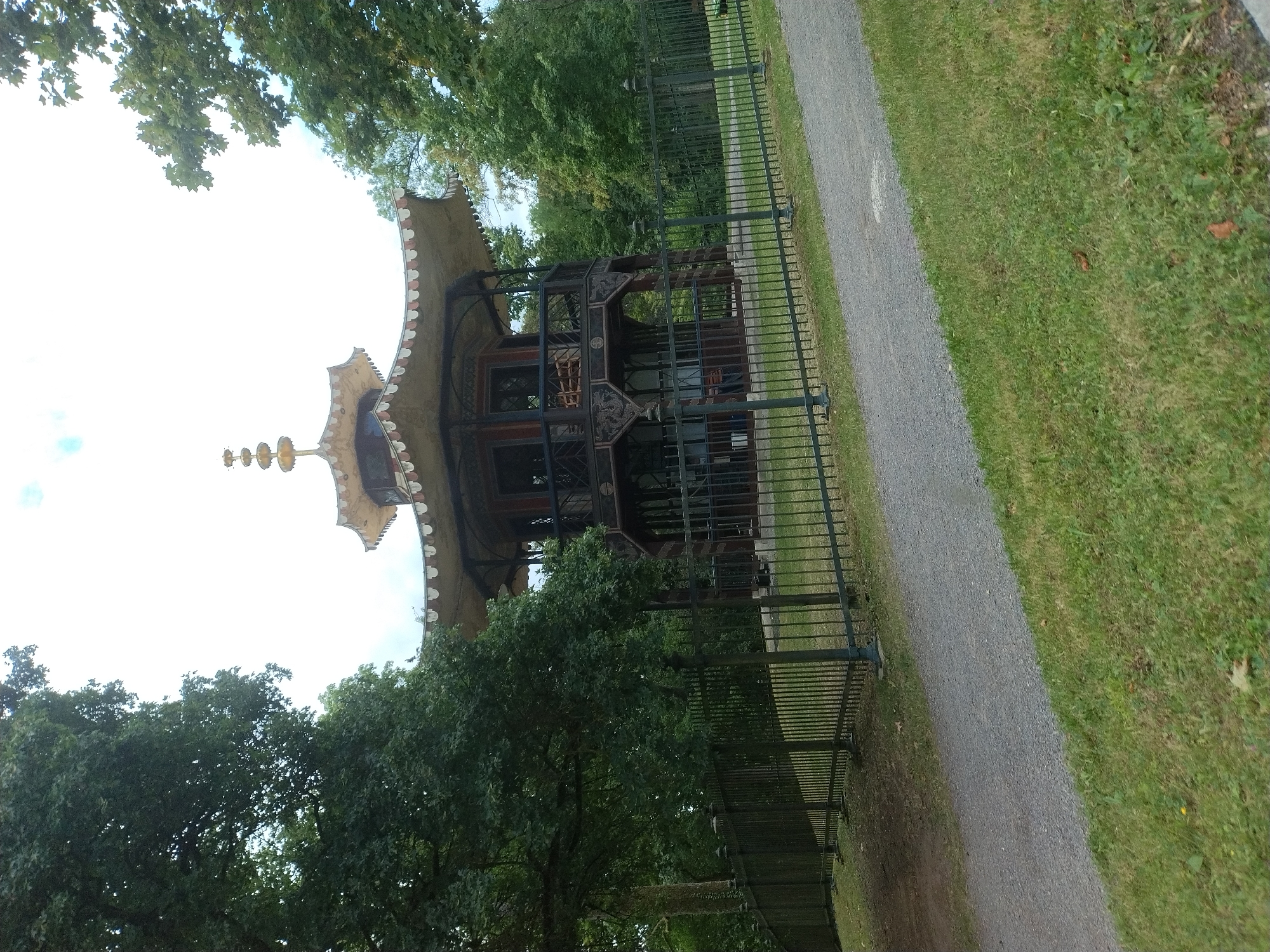 The image shows a Chinese pagoda in a park-like setting. The pagoda is a multi-tiered structure with dark wood framing and a light-colored, ornate roof. It appears to be a building, possibly a pavilion or gazebo, with open sides on the upper level revealing wooden benches inside. The pagoda is surrounded by lush green trees and a black metal fence encloses a grassy area in front of it. A gravel path runs diagonally across the foreground, with more grass to the right. The sky is overcast. The pagoda is located in the "Walhalla" park in Donaustauf, Germany.