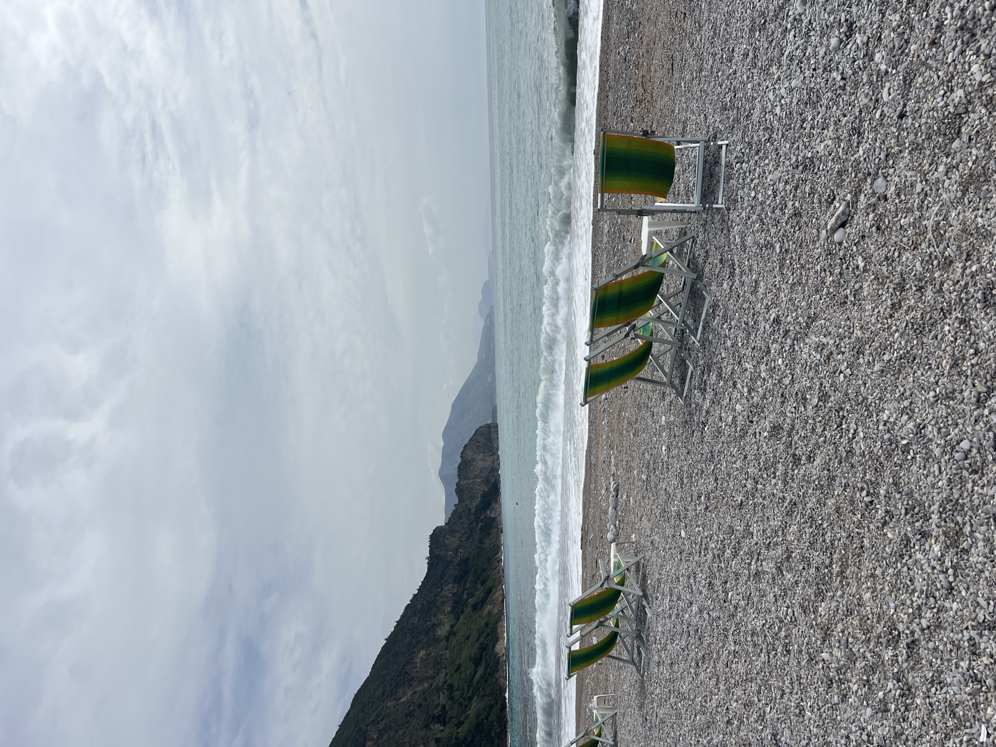 The image shows a beach scene in Prijevor, Montenegro, featuring a row of unoccupied beach chairs with green and yellow striped fabric on a pebble beach. The sea is rough with white waves crashing onto the shore. In the background, a steep, rocky cliff covered with green vegetation rises up from the shoreline. The sky is overcast with grey clouds.