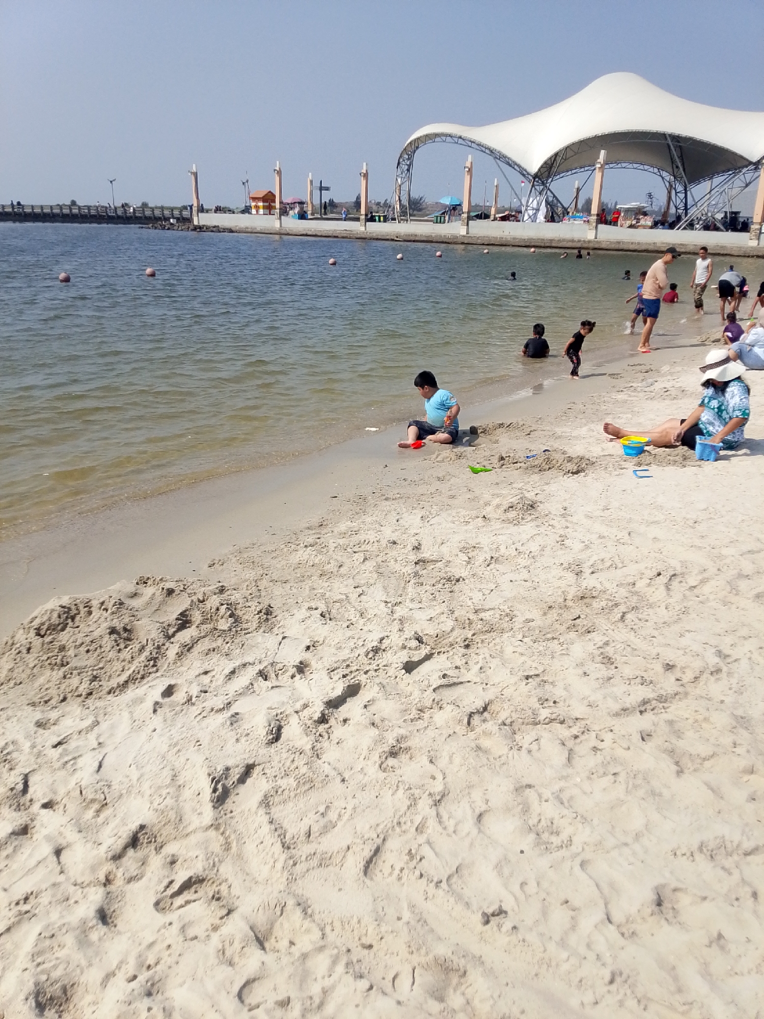 The image depicts a beach scene at Ancol Beach City, North Jakarta, Indonesia, on a clear, sunny day. The foreground is dominated by a sandy beach with visible footprints and sand formations. Several people are present, engaged in various beach activities.
In the mid-ground, a young boy in a blue shirt is sitting on the sand playing with red and green sand toys. To his right, a woman wearing a straw hat and a patterned shirt is sitting on the sand with two buckets, one blue and yellow, and another blue one. She appears to be interacting with the sand. Further into the water, several children and adults are wading and playing.
The water's edge is clearly defined, with gentle waves lapping the shore. In the background, a long pier extends into the water, lined with lamps and some structures. Beyond the pier, a large, white, tent-like structure with a metal frame is visible, characteristic of the entertainment facilities in the Ancol area. The sky is a clear, pale blue. The overall atmosphere is one of leisure and recreation.