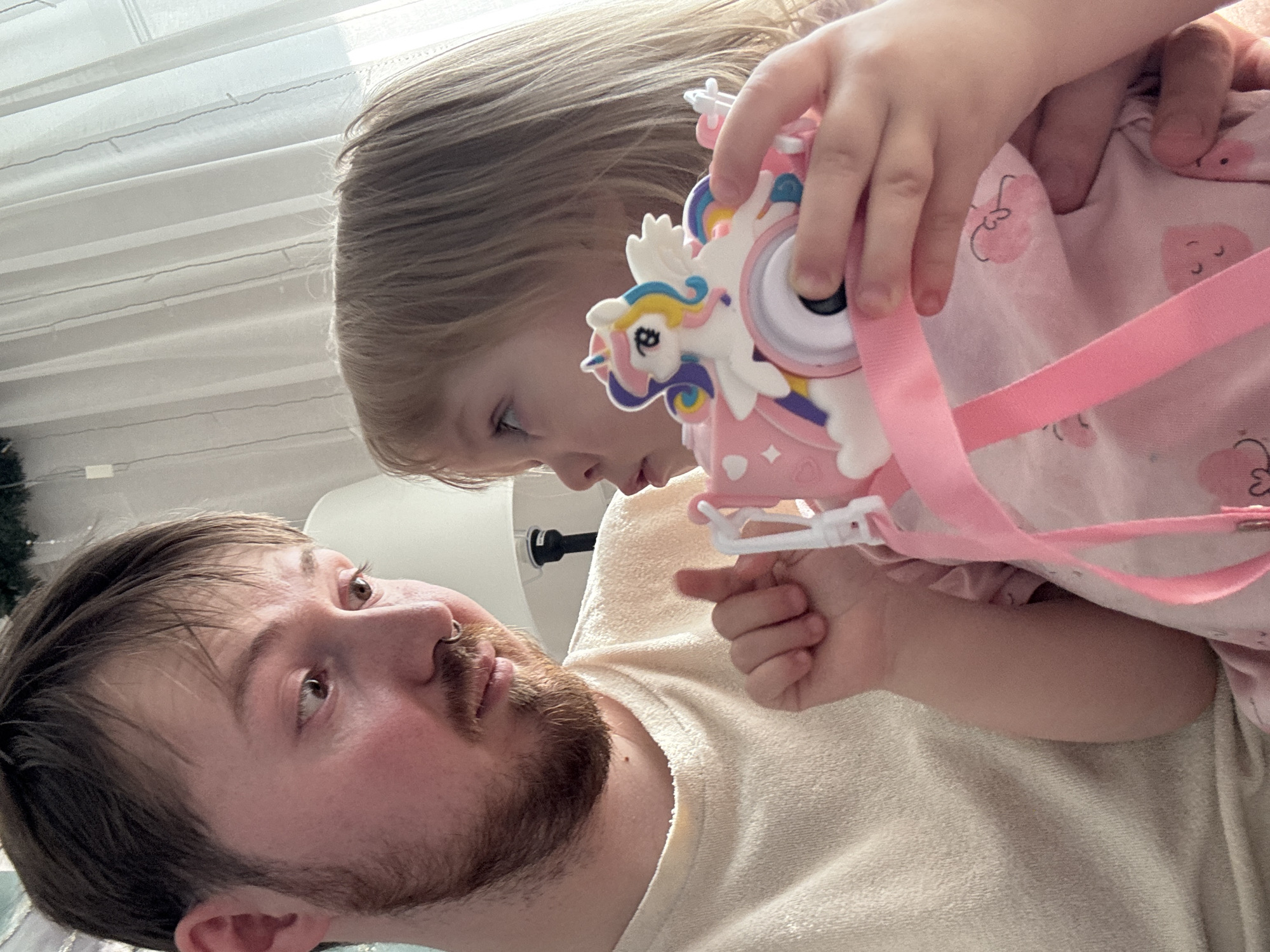 An indoor, eye-level shot captures a tender interaction between an adult man and a young child. The man, with a neatly trimmed beard, dark hair, and a septum piercing, is positioned in the lower left of the frame, looking up and slightly to his right with a gentle, engaged expression. He is wearing a light-colored, possibly terry cloth, top. Above him, a young child with blonde hair is holding a white and pink unicorn-themed toy, which appears to be a small camera or bag, complete with a pink strap. The child, dressed in a pink garment adorned with small patterns resembling hearts or clouds, focuses intently on the toy and her interaction with the man.
The background is softly lit by natural light, indicating daytime. Horizontal white blinds or curtains are visible, creating a bright but diffused setting. A subtle hint of green foliage, possibly from a houseplant or holiday decoration, can be seen on the far left. The composition highlights the close bond and shared moment between the two subjects, conveying a sense of warmth and affectionate play within a domestic setting in Chaikovsky, Russia. No visible text is present in the image.