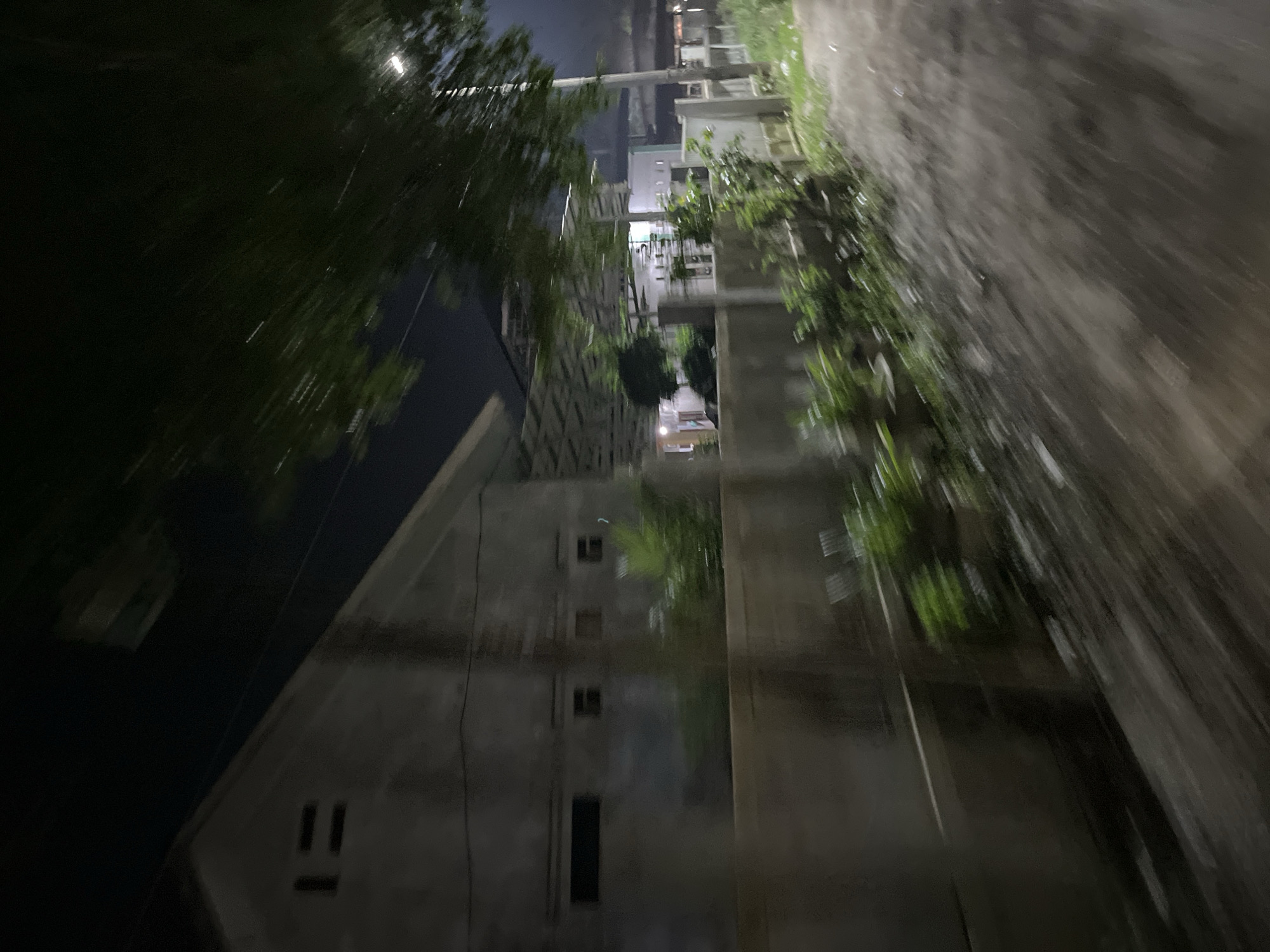 This image depicts a street scene at night in what appears to be an urban or suburban area. The perspective is low and angled, suggesting it was taken from the ground or a low vehicle, possibly while in motion due to the motion blur.
The scene is dominated by buildings, some of which are multi-story structures with visible windows. Trees and foliage are present, blurring into the background and foreground, indicating a possibly damp or rainy environment as streaks of light suggest water in the air. The lighting is dim, consistent with nighttime, with some artificial lights from the buildings casting a glow. The overall mood is somewhat mysterious due to the darkness and blur. The ground appears to be a paved surface, possibly a road or walkway. A notable detail is the presence of what looks like a simple smiley face drawn on the side of one of the buildings, formed by two vertical lines and a horizontal line.
The location context provided is "Kesehatanbandar, Indonesia." Given the architectural style and vegetation, this is plausible. The time of day is clearly night, and the weather might be rainy or recently rained due to the streaks of light and blurry appearance of the trees. There is no visible text in the image other than the rudimentary smiley face.