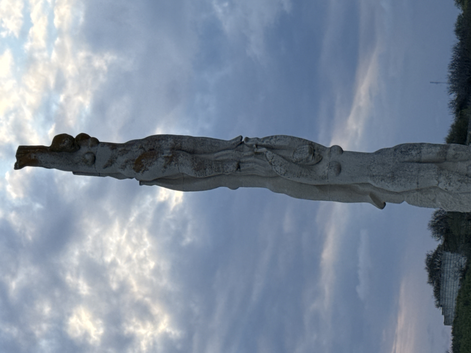 The image shows a weathered concrete monument in Balgarevo, Bulgaria, set against a cloudy sky. The monument's horizontal, elongated design features stylized shapes suggestive of entwined figures or animals. The structure exhibits signs of age and wear. To the right, there is vegetation and a low concrete structure, possibly part of a building or a retaining wall.