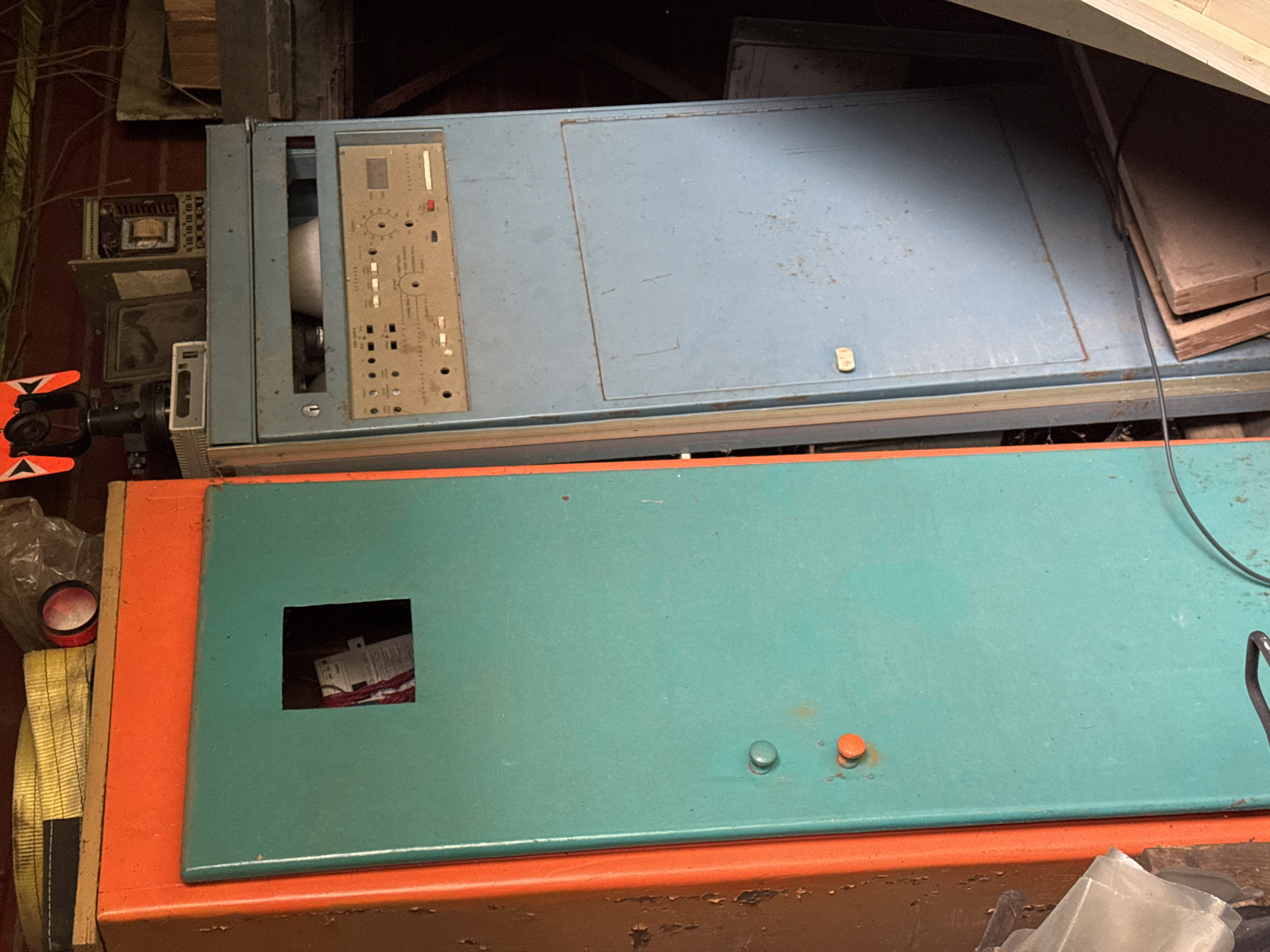 The image depicts a cluttered indoor space, possibly a workshop or storage area. Two large, retro-looking electronic cabinets dominate the foreground. The cabinet on the left is orange and teal, with a square opening revealing wires and what appears to be labeling inside. The cabinet on the right is blue, with a control panel featuring numerous buttons, dials, and a small screen. Other objects visible include what looks like a surveying instrument on top of the blue cabinet, a yellow strap, and some wooden planks in the background. The lighting suggests it is daytime, but the environment appears somewhat dim. There are no people or animals in the image. The location of Naro-Fominsk, Russia, is provided as context, but the image itself does not contain any visual cues to confirm this specific location.