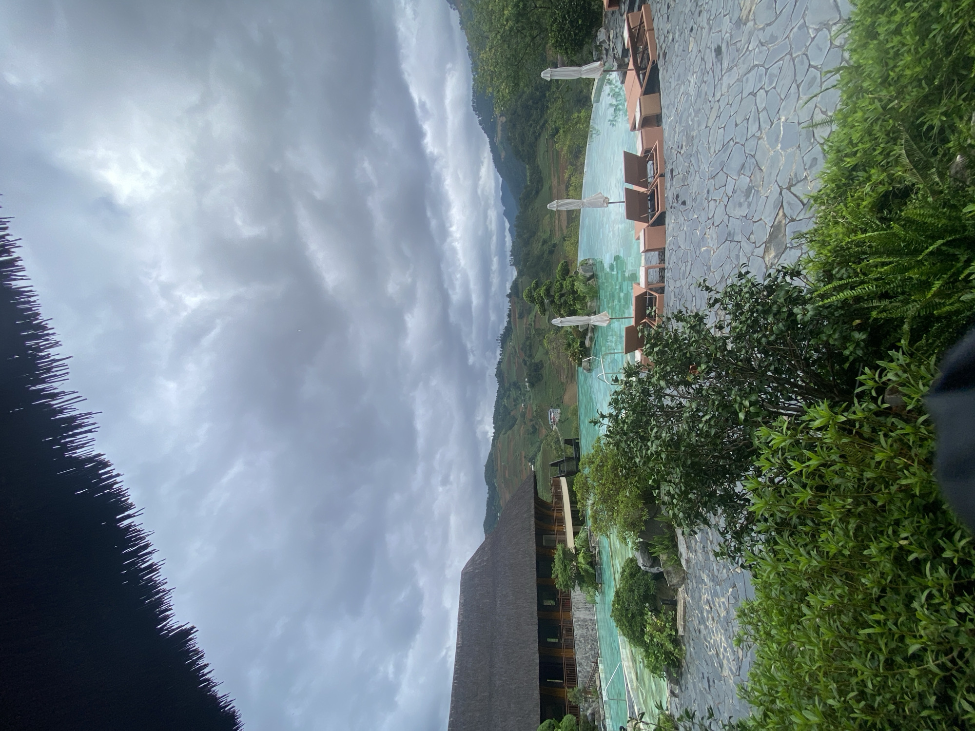 The image depicts an outdoor recreational area in Púng Luông, Vietnam, under an overcast sky. A large, irregularly shaped swimming pool with light turquoise water occupies the midground, featuring small islands with green foliage and rocks, including a sculpted miniature tree. Several brown lounge chairs with small side tables and white parasols (some open, some closed) are arranged around the pool's edge, which is paved with dark grey flagstones. To the left of the pool stands a multi-story building with a dark, possibly thatched roof and light-colored wooden walls, featuring balconies overlooking the pool and landscape. A wooden staircase is visible on the right side of the building. In the background, a series of green, terraced mountains and hills stretch towards the horizon under a heavily clouded, grey sky with some brighter patches. The foreground is partially obscured by the edge of a dark, thatched roof at the top and dense green bushes and foliage at the bottom. No distinct individuals are visible.