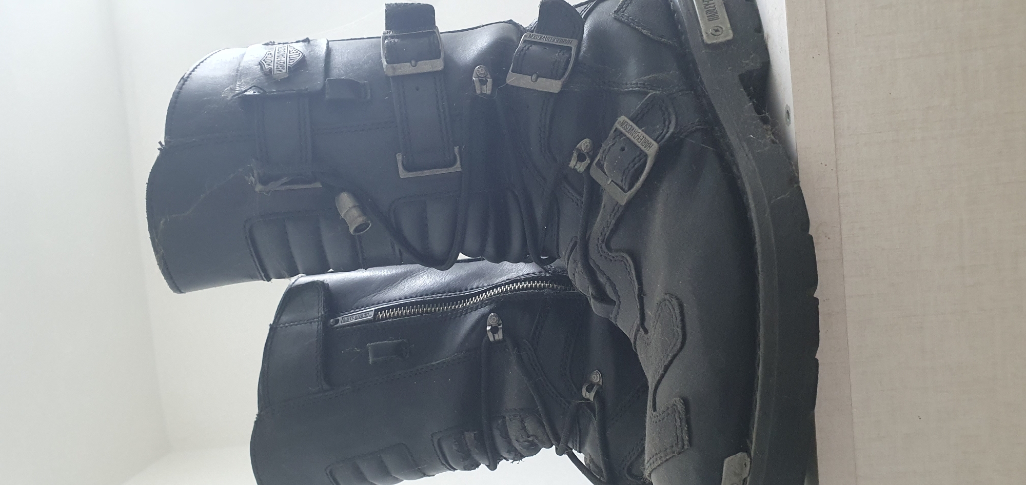 A pair of black leather Harley-Davidson motorcycle boots are placed on a white, textured surface. The boots are positioned side-by-side, with the left boot slightly in front of the right. Both boots feature multiple buckles and straps, as well as a zipper closure on the left boot. The Harley-Davidson logo is visible on several parts of the boots. The boots appear to be worn and show signs of use, including scuff marks and creasing. The background is a plain white wall. The image was taken indoors, likely in a residential setting in Bat Yam, Israel, based on the implied context of the request.