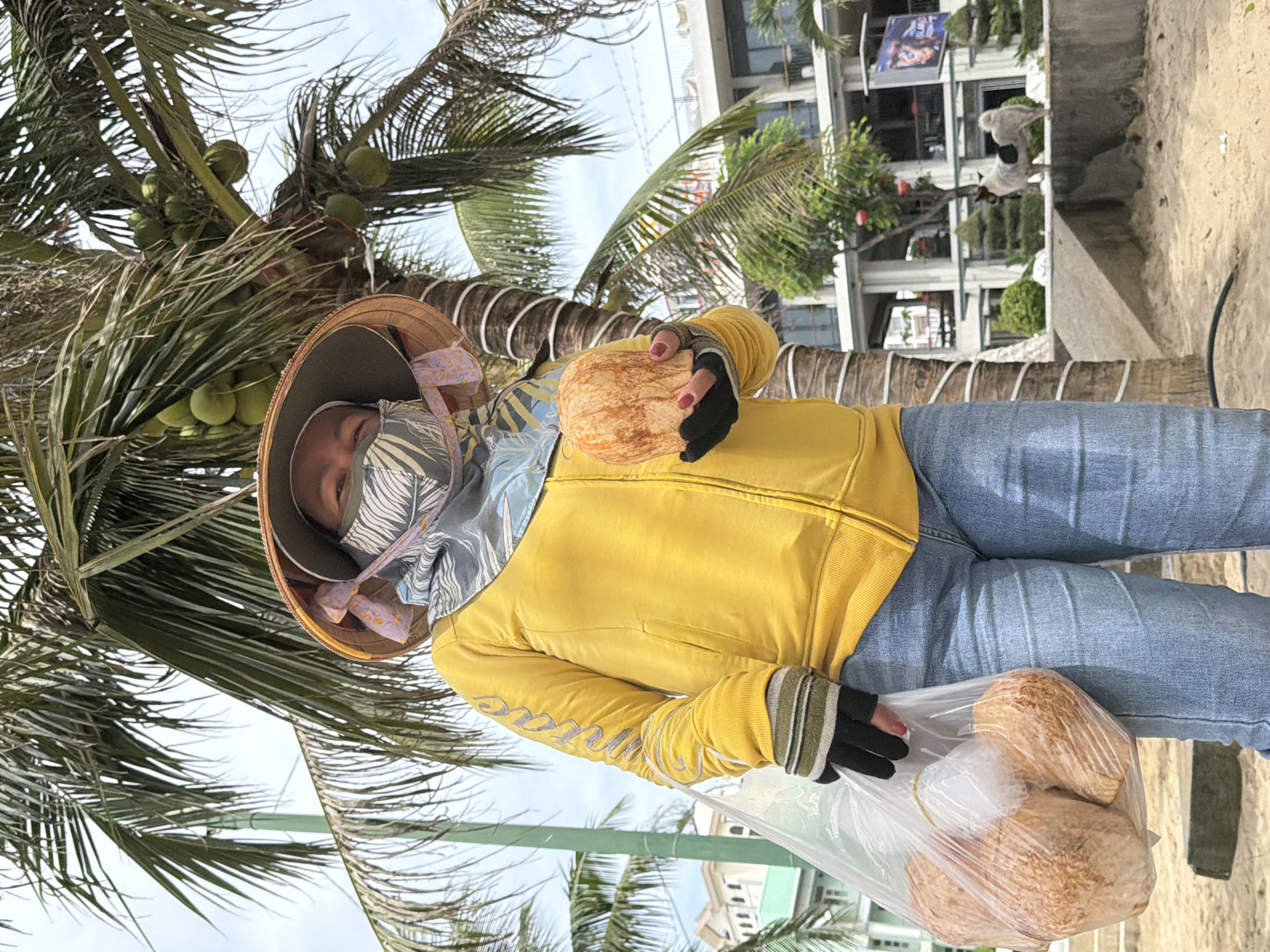 A person stands on a sandy beach in Nha Trang, Vietnam, holding a large coconut in their right hand and a plastic bag filled with coconuts in their left. The person is wearing a yellow zip-up jacket with a patterned mask and a conical hat. Behind them, a palm tree with coconuts is visible, along with a building in the background. The weather appears to be overcast.
